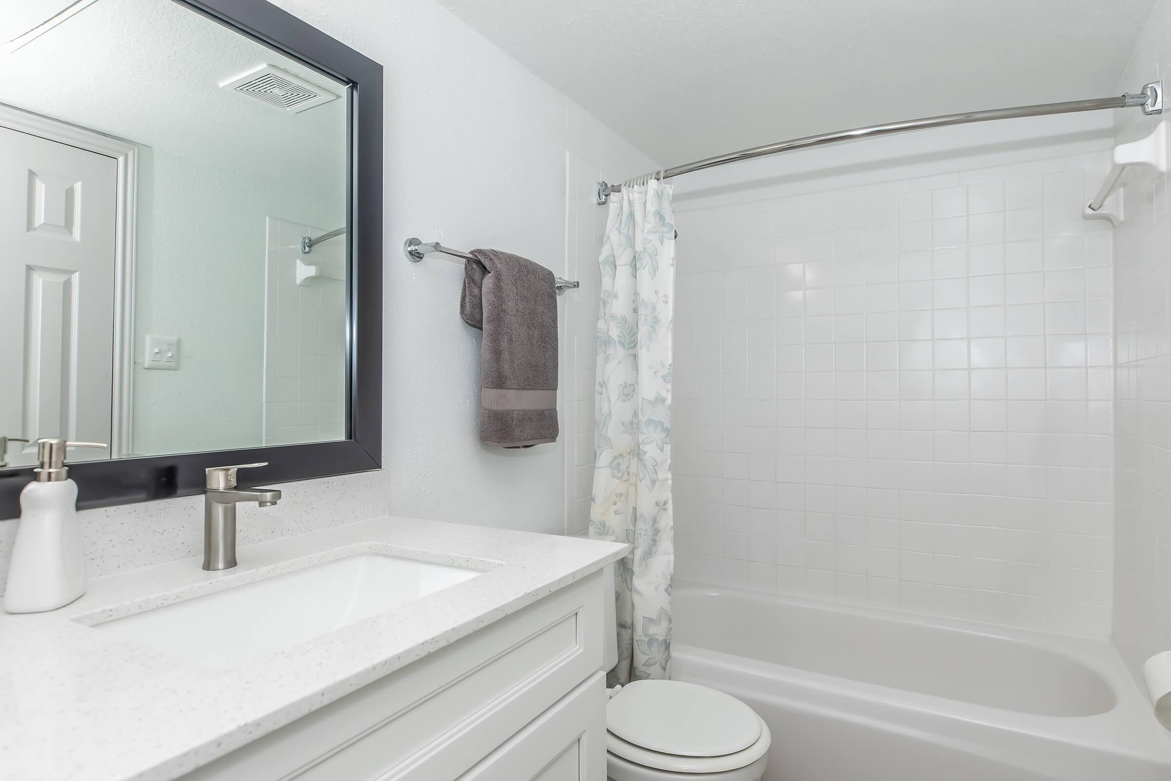 A clean and modern bathroom featuring a white bathtub, a shower with a floral curtain, a sleek sink with a modern faucet, and a large mirror above the sink. Soft gray towels hang on a towel rack, and the walls are light-colored, creating a bright and airy atmosphere.