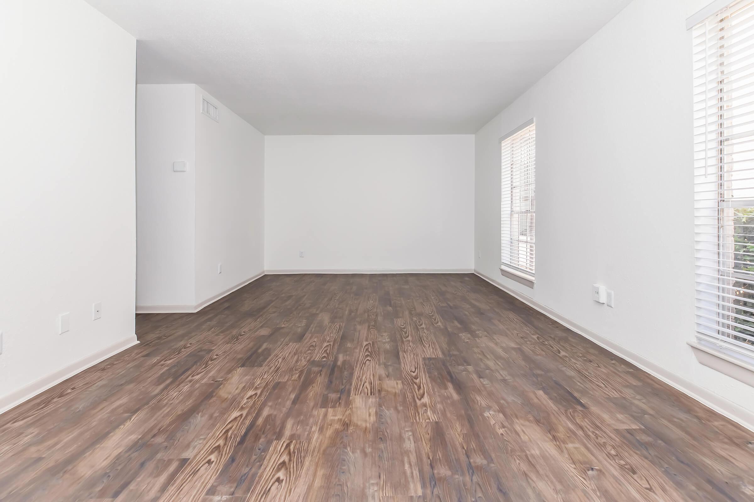 A spacious, empty living room with light-colored walls and large windows featuring blinds. The floor is covered in dark wooden planks, creating a warm contrast. The overall atmosphere is bright and airy, with natural light illuminating the space. No furniture or decorations are present.