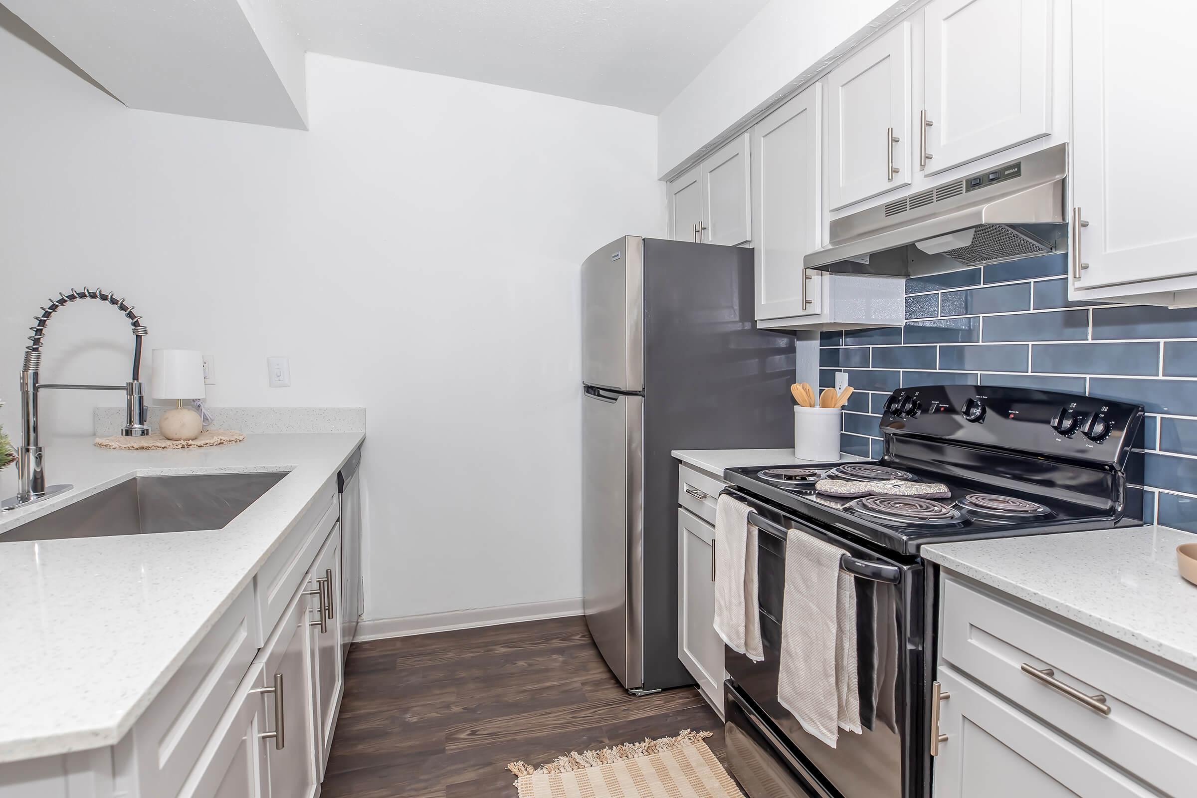 Modern kitchen with white cabinets, a stainless steel refrigerator, a black stove with oven, and a sleek sink. The backsplash features blue tiles, and the countertops are light-colored. Kitchen utensils are neatly arranged, and a beige rug lies on the floor, creating a clean and inviting atmosphere.