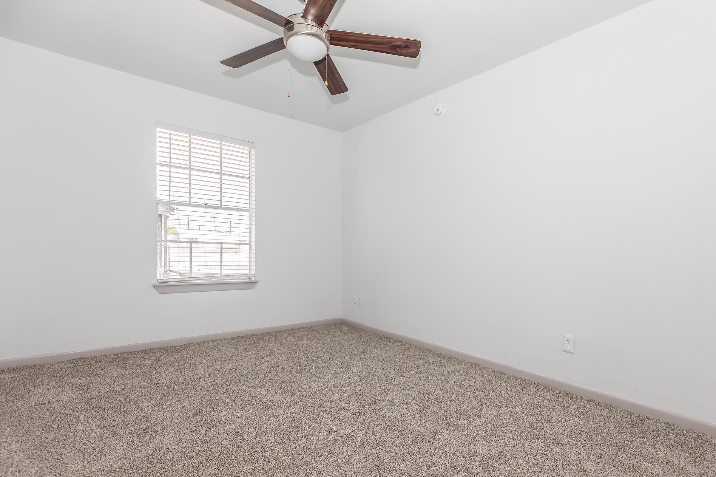 A bright, empty room with light-colored walls and beige carpet. It features a ceiling fan with wooden blades and a window with horizontal blinds, allowing natural light to enter. The room is spacious and uncluttered, creating a minimalist atmosphere.