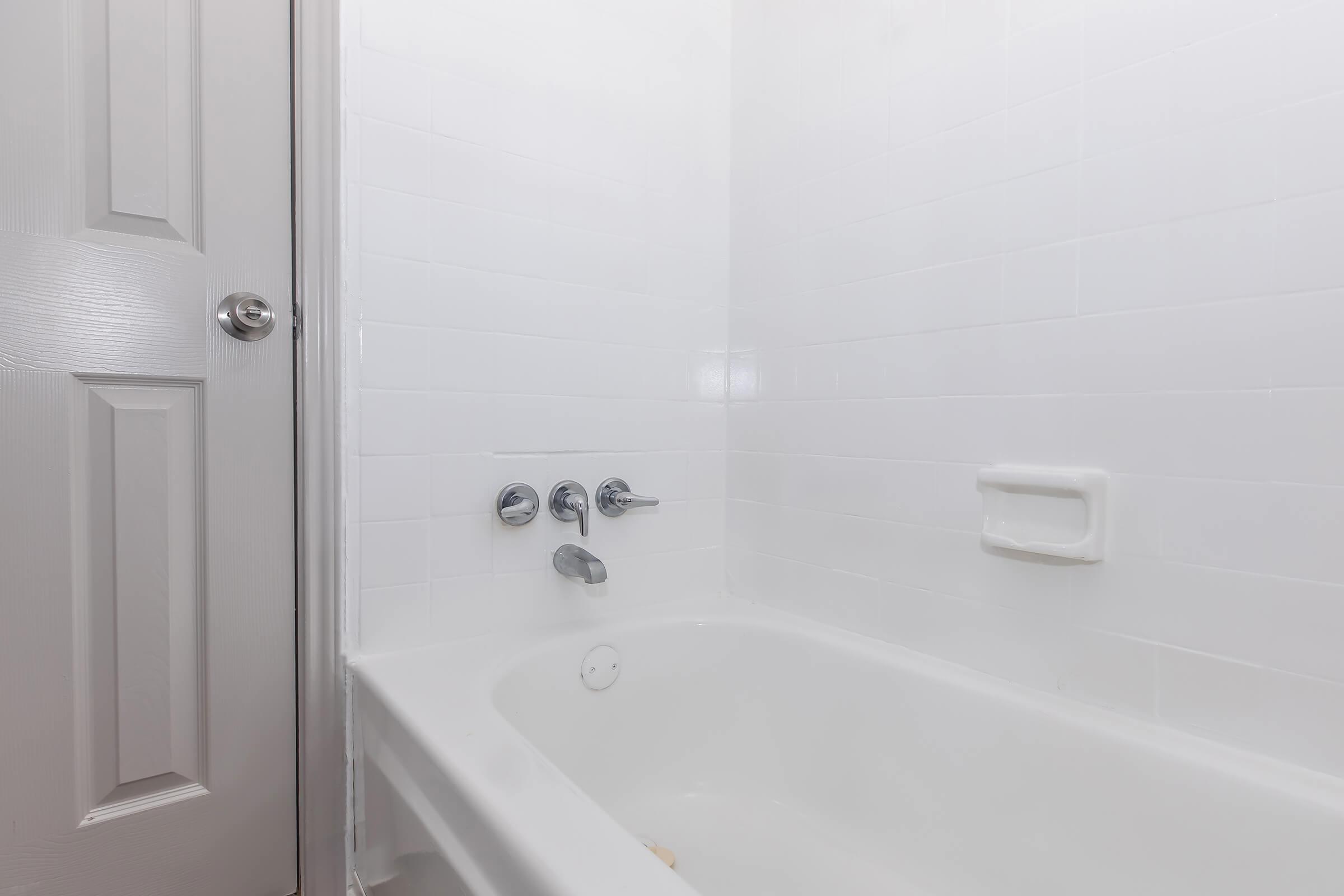 A clean, empty white bathtub with modern fixtures, set against a white tiled wall. A closed door is visible in the background, also white, contributing to the minimalist aesthetic of the bathroom. The overall atmosphere suggests a bright and airy space.