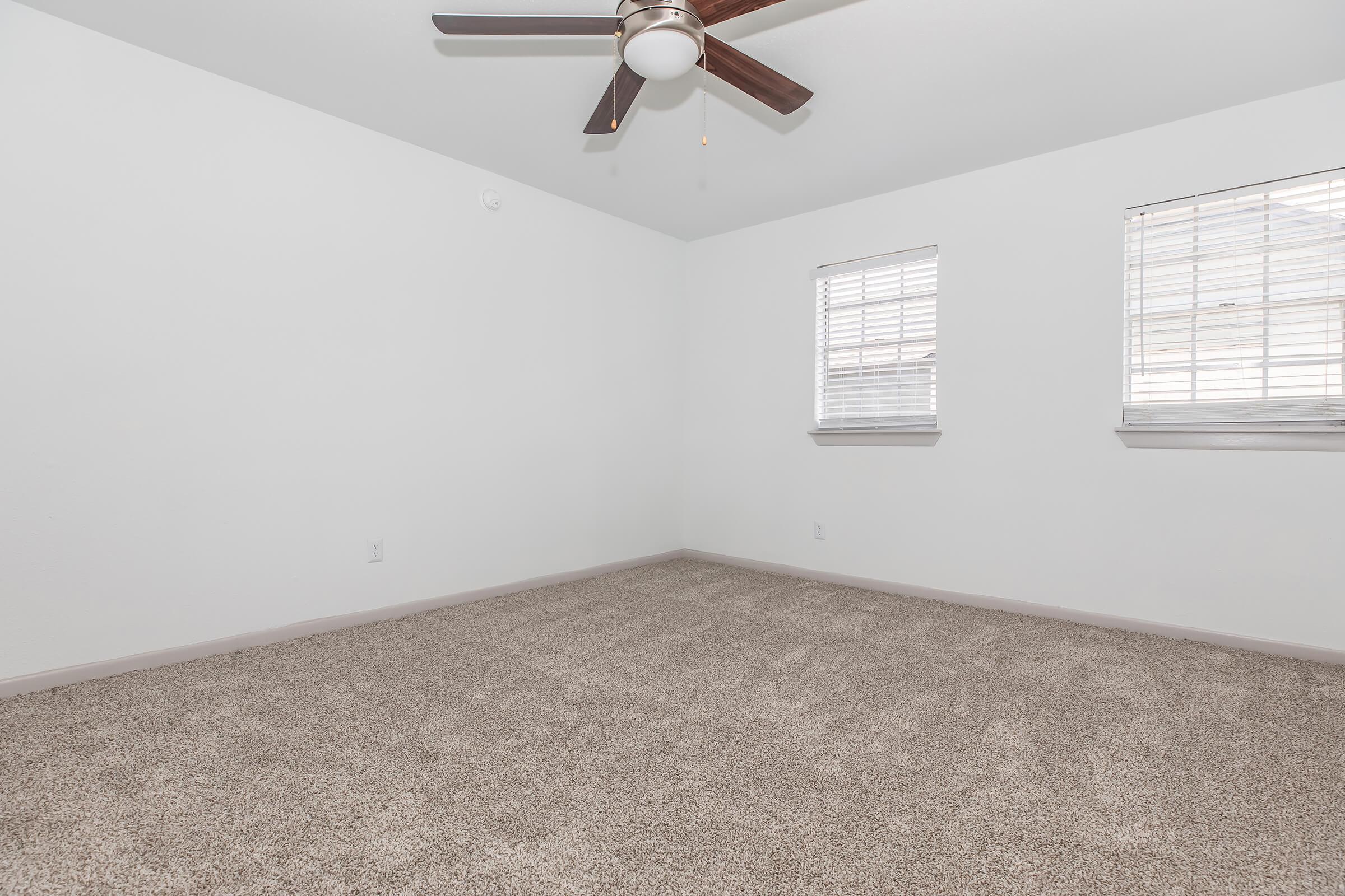 A spacious, empty room with light-colored walls and a ceiling fan. The floor is covered in plush, beige carpet. There are two windows with blinds allowing natural light to enter, creating a bright and airy atmosphere.