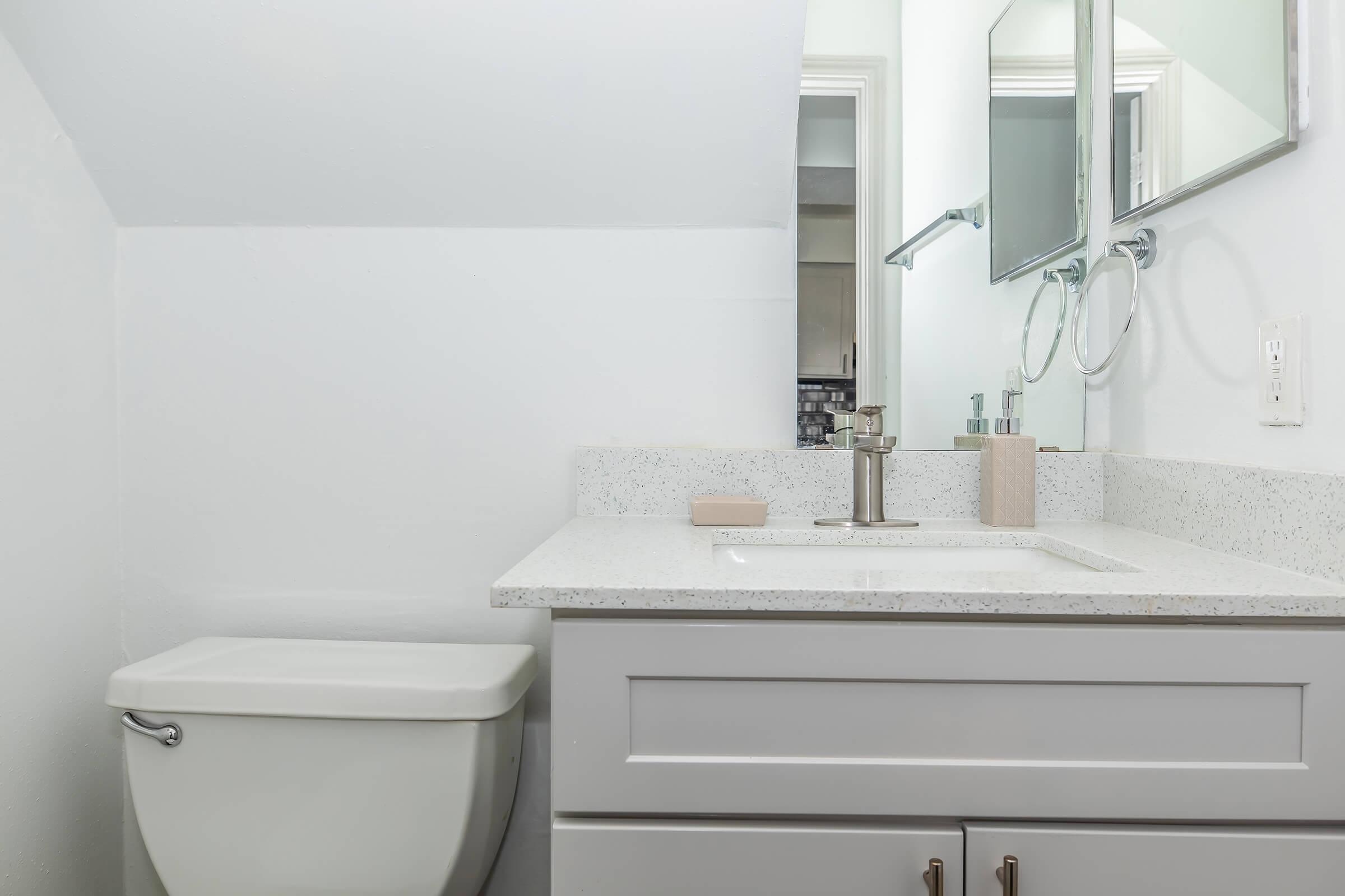 A clean, modern bathroom featuring a white wall and a sloped ceiling. The space includes a white toilet, a sleek sink with a light-colored countertop, and a mirror above it. There are minimal decorative elements, creating a tidy and spacious feel.