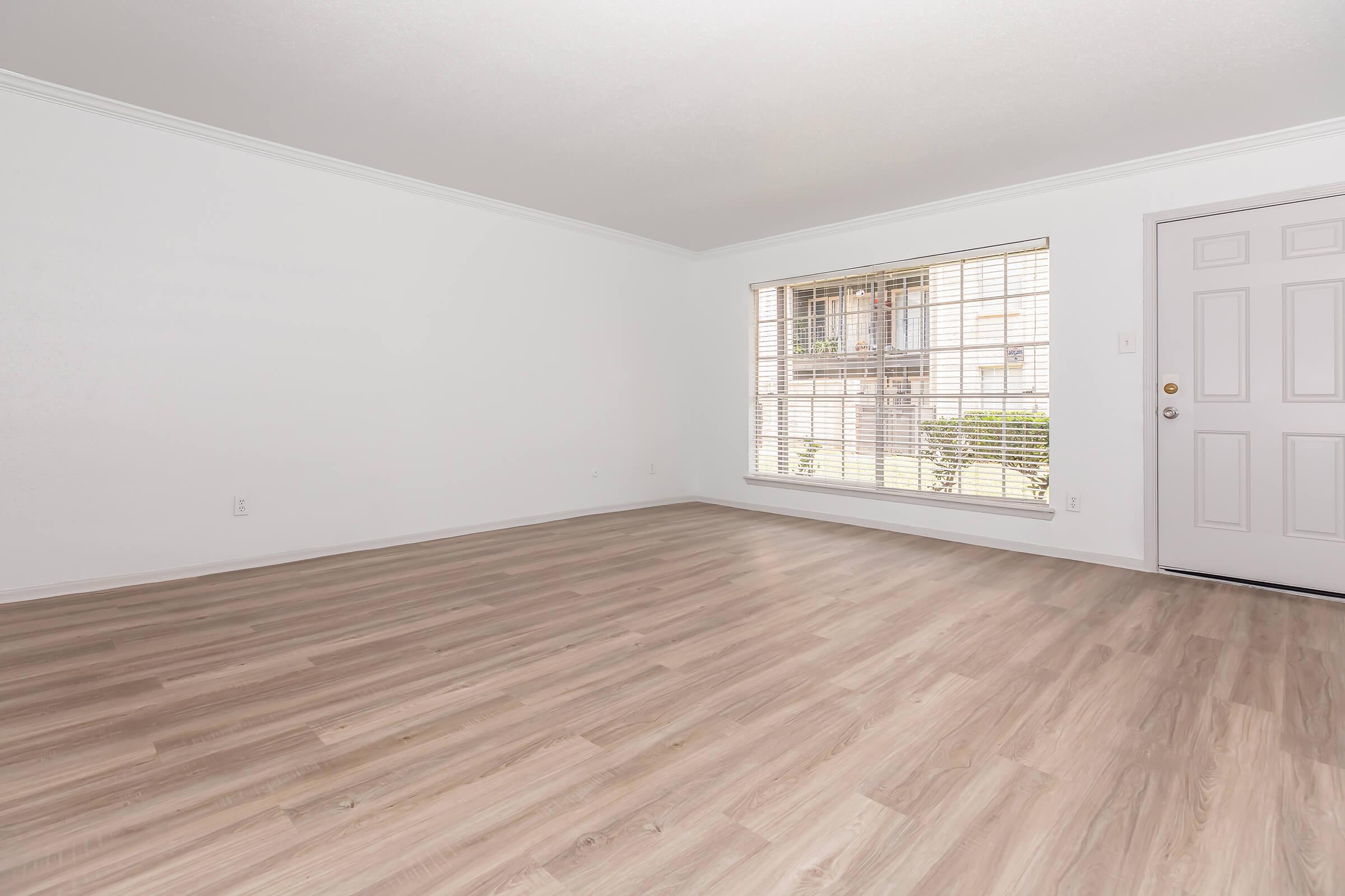 A spacious, empty room with light-colored wood laminate flooring. The walls are white, and there is a large window with blinds allowing natural light to enter. A front door is visible on the right side, enhancing the room's open and airy feel.