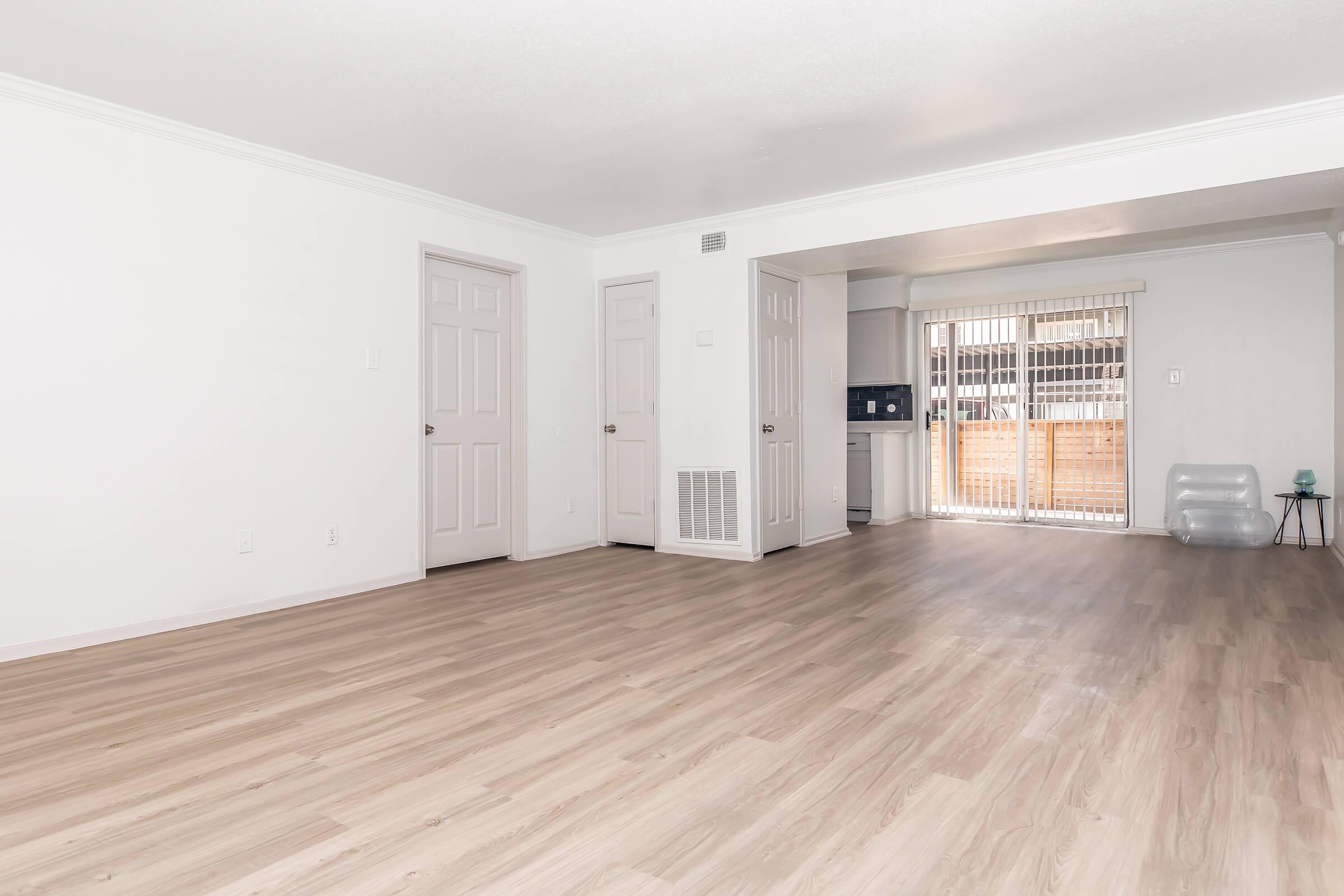 A spacious room with light-colored walls and laminate flooring. There are two closed doors on the left, and large sliding glass doors leading to an outdoor area on the right. The kitchen is partially visible in the background, showcasing a modern, open layout with natural light.