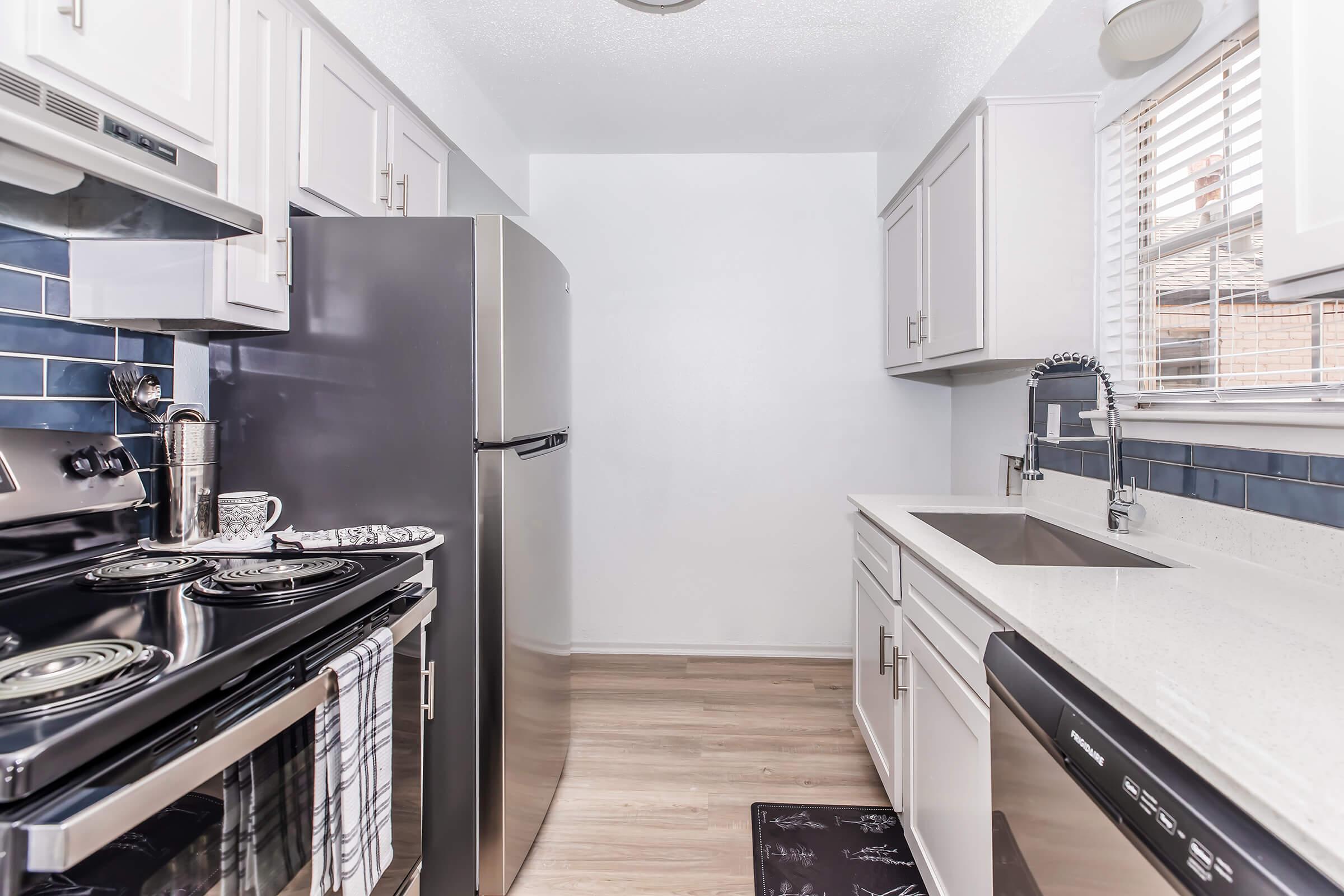 Modern kitchen featuring stainless steel appliances, including a refrigerator and oven. There is a sleek countertop with a sink and a dishwasher, along with light cabinetry and blue tiled backsplash. The space is well-lit with natural light coming from a window.
