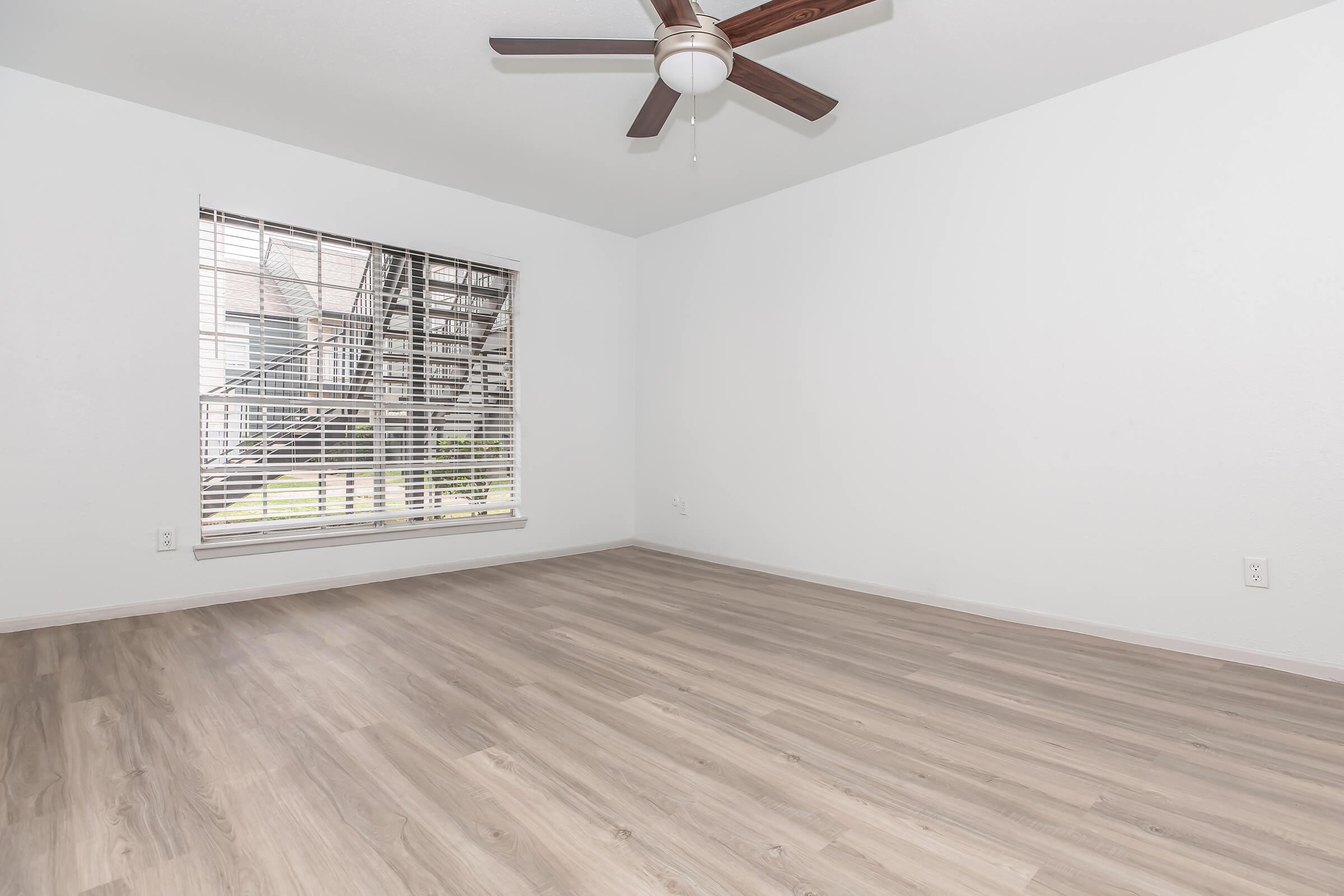 A clean, empty room featuring light wooden flooring, a ceiling fan, white walls, and a large window with blinds. The view outside includes a staircase and greenery. The space appears bright and spacious, with no furniture present.