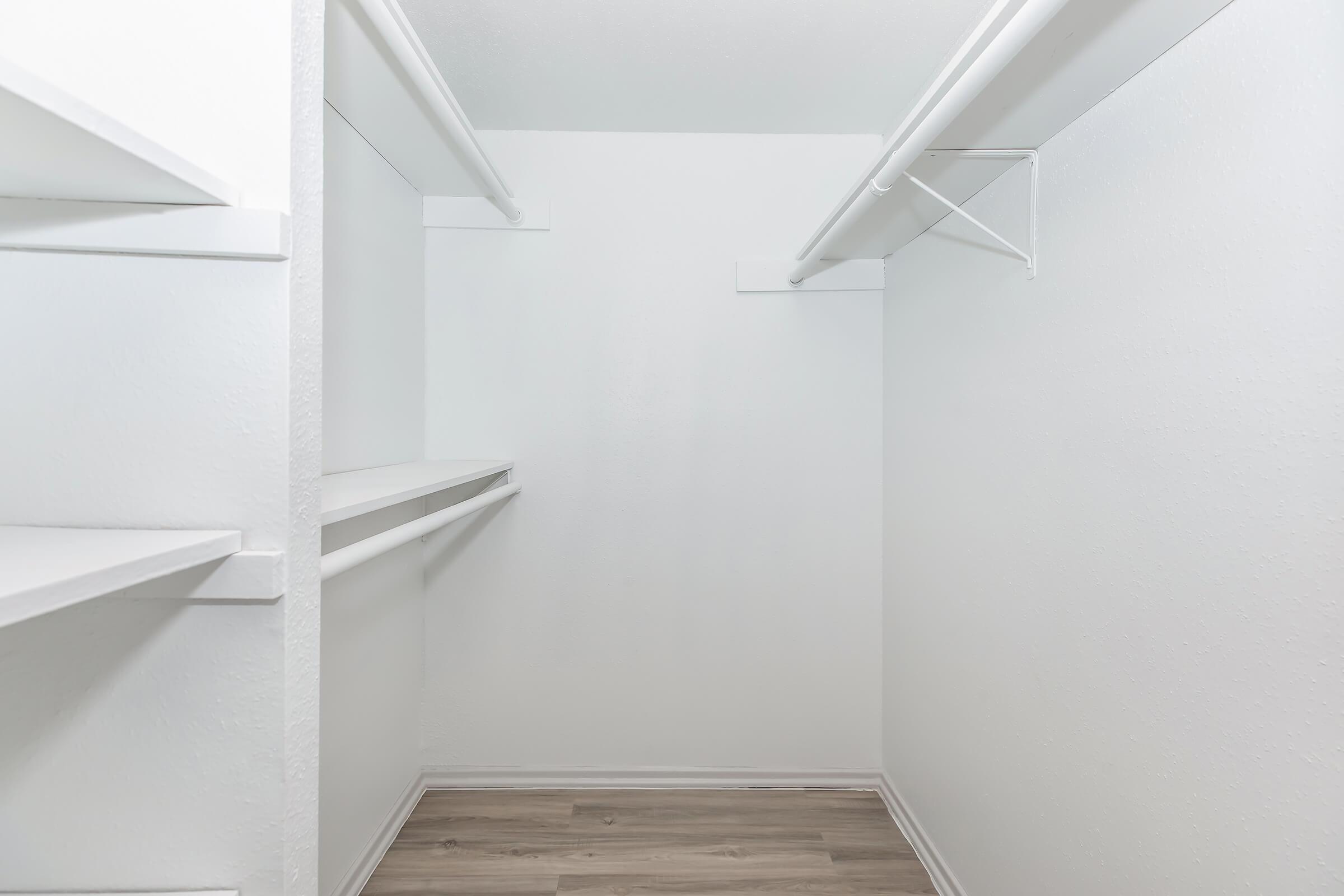 Empty walk-in closet featuring white walls and wooden flooring. The closet includes shelves on one side and a hanging rod on the opposite wall, providing ample storage space. The environment appears clean and uncluttered, suitable for organizing clothing and accessories.