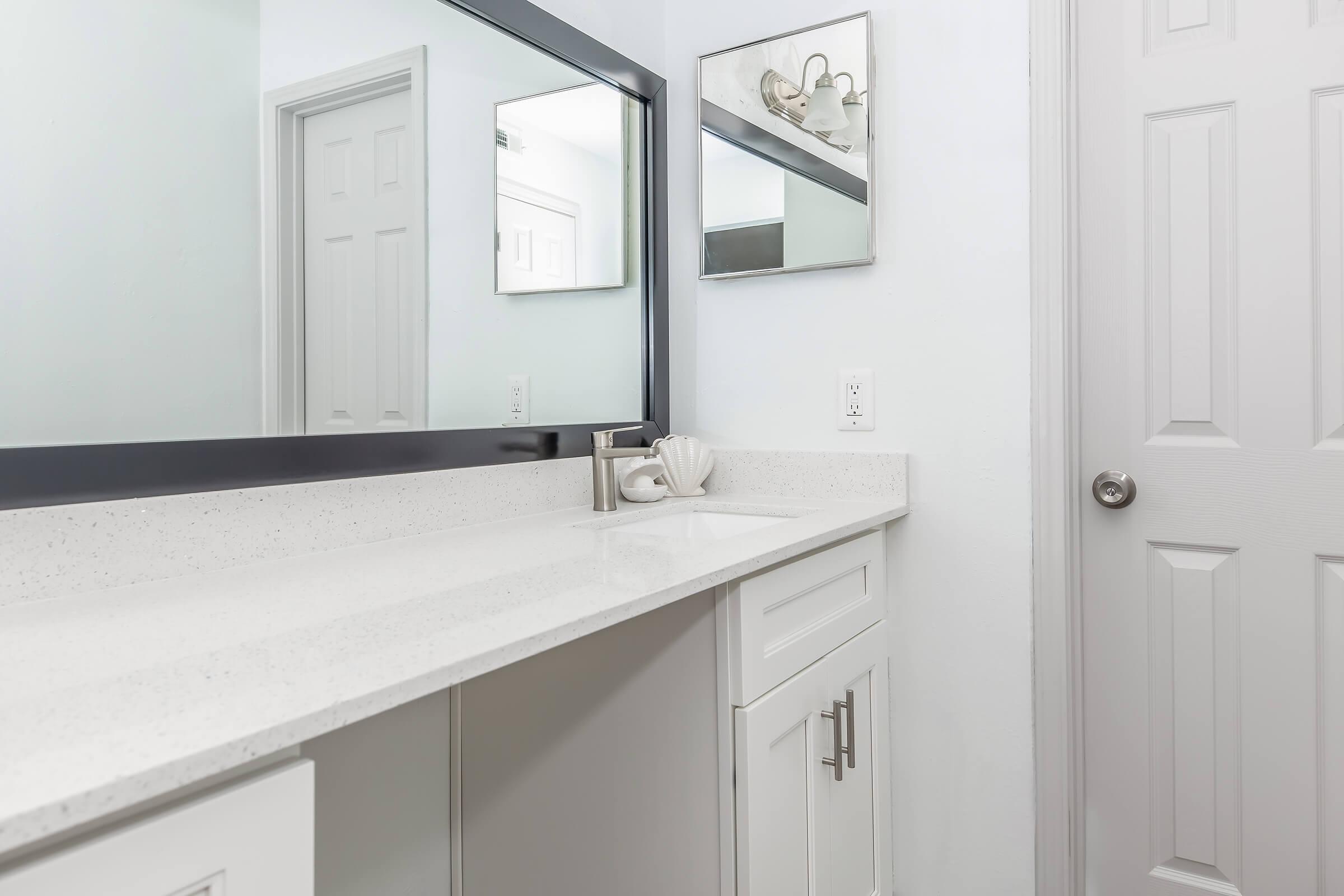 A modern bathroom featuring a white countertop and a large mirror. The cabinetry is sleek and white, with a minimalist design. A subtle decorative item is placed next to a faucet, and there is a closed door with a round doorknob in the background, completing the clean and contemporary look.