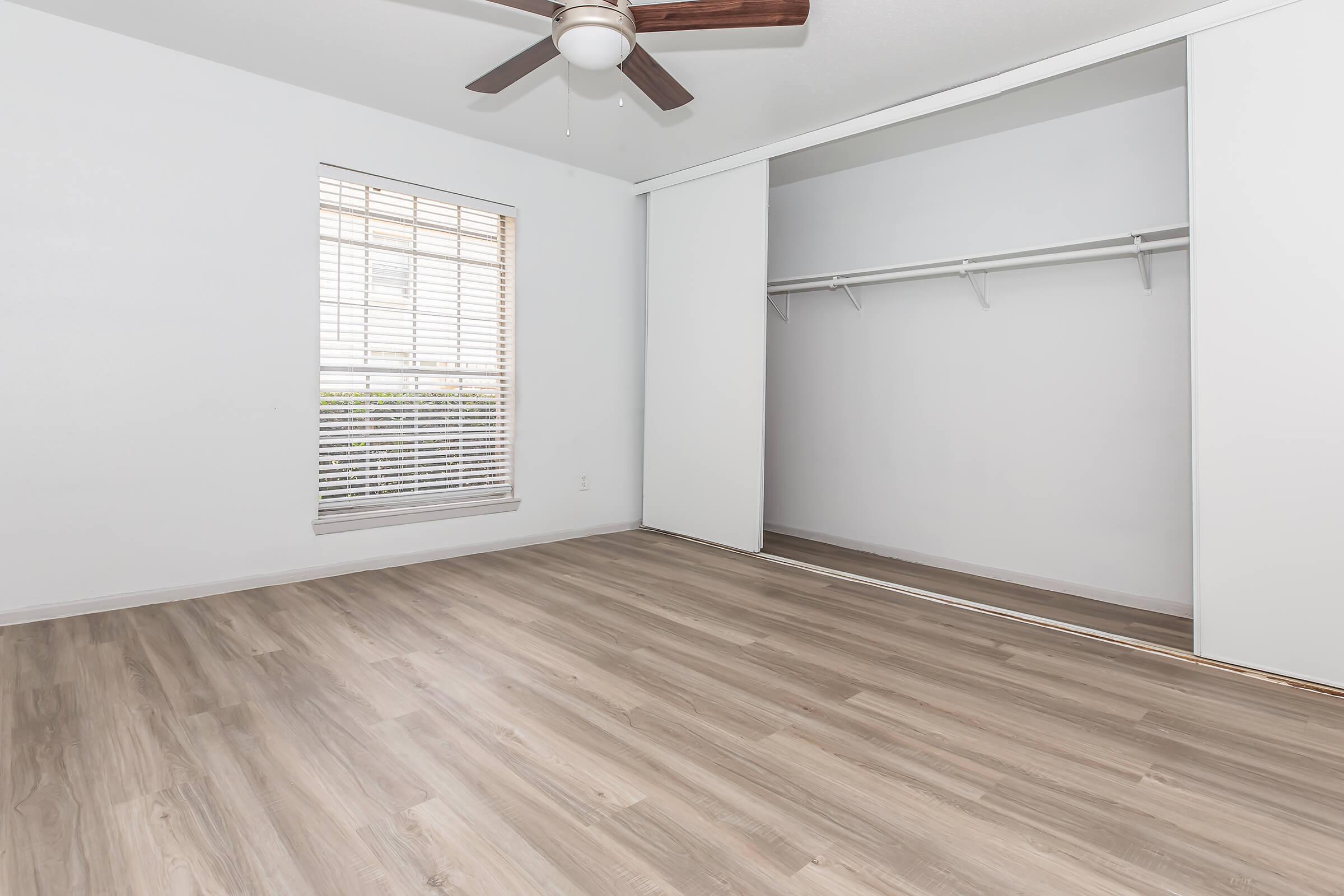 A bright, empty room with light wood flooring, a ceiling fan, and a window featuring horizontal blinds. There's a sliding closet door along one wall, and the space has white walls and natural light coming through the window.