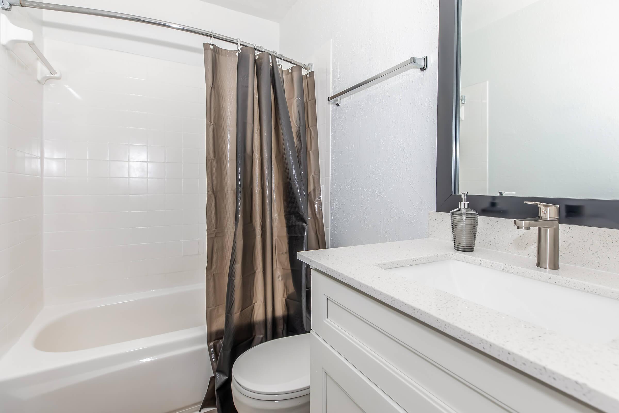 A modern bathroom featuring a bathtub with a shower curtain, a white sink with a countertop, and a large mirror. The decor includes a sleek faucet and a decorative piece on the sink. The walls are painted white, contributing to a clean and bright atmosphere.