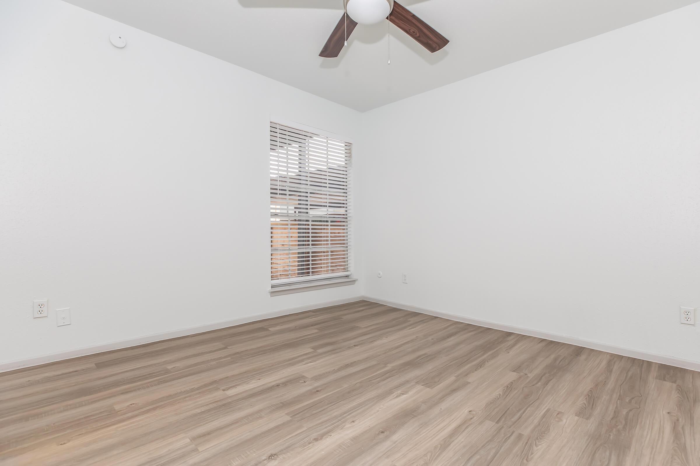 A clean, empty room with light-colored walls, a ceiling fan with wooden blades, and a window with horizontal blinds. The floor is a light wood laminate, creating a bright and airy atmosphere. The room has minimal decor, making it suitable for various uses.