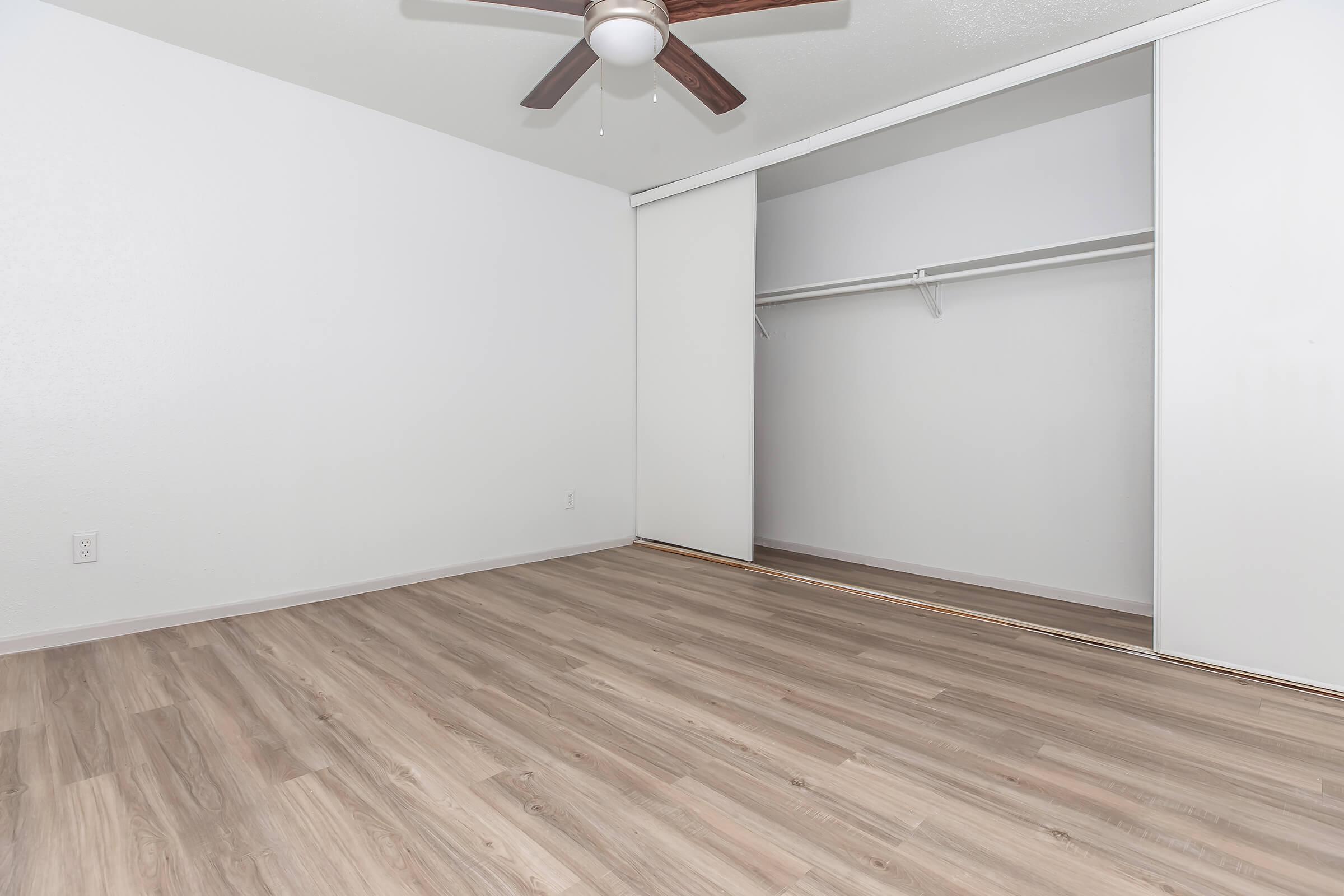 A spacious, empty room featuring light-colored walls and a wood laminate floor. It has a ceiling fan and a sliding closet with a hanging rod. Natural light illuminates the uncluttered space, creating a bright and airy atmosphere.