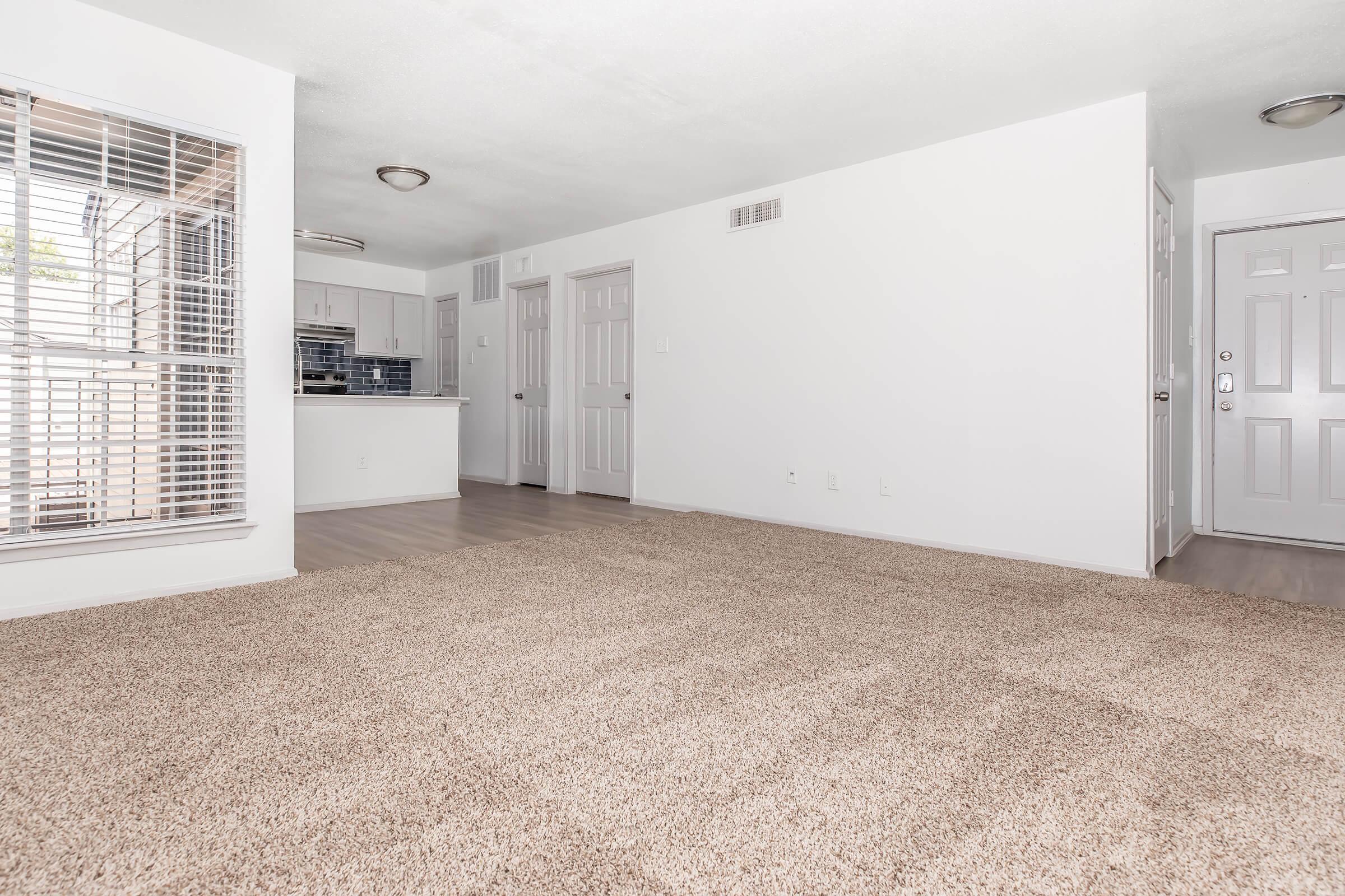 A spacious, vacant living room with light beige carpeting, large windows letting in natural light, and a partially visible kitchen area in the background. The walls are painted white and the doors are light-colored. The overall ambiance is bright and clean, suitable for various interior design styles.