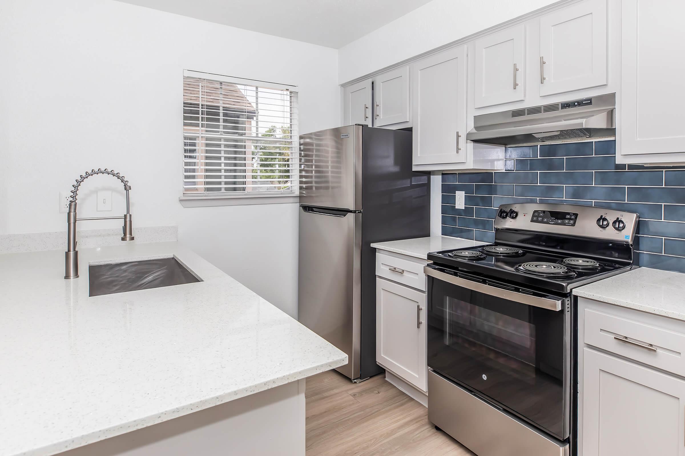 Modern kitchen featuring stainless steel appliances, including a refrigerator and an electric stove with a sleek design. The countertops are light-colored with a shiny finish, complemented by blue backsplash tiles. A window allows natural light to enter, and there is a stainless steel sink with a modern faucet.