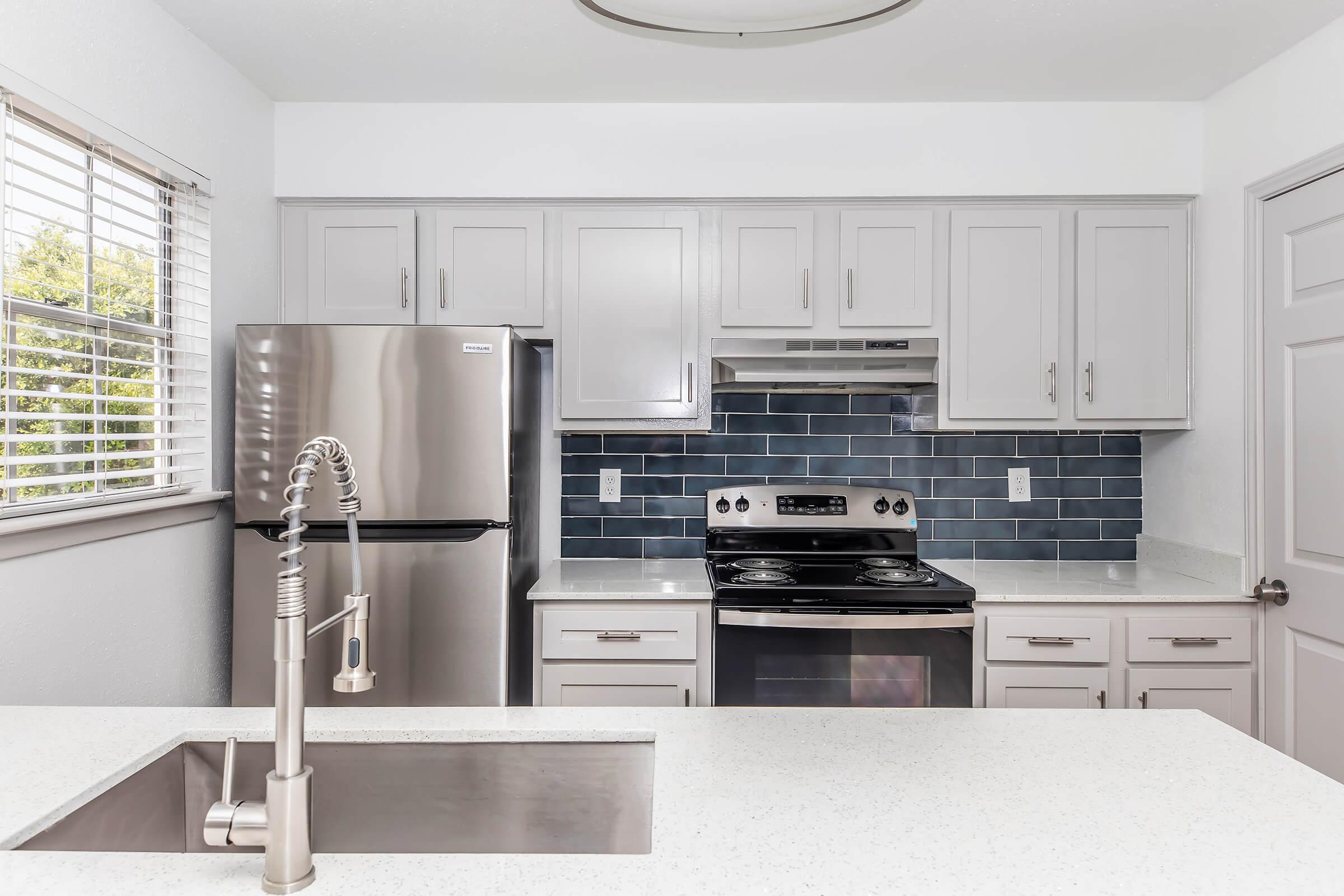 Modern kitchen featuring stainless steel appliances including a refrigerator and oven. The cabinetry is light gray, complemented by a dark blue tile backsplash. A spacious white countertop with an undermount sink is visible, along with a window that lets in natural light.