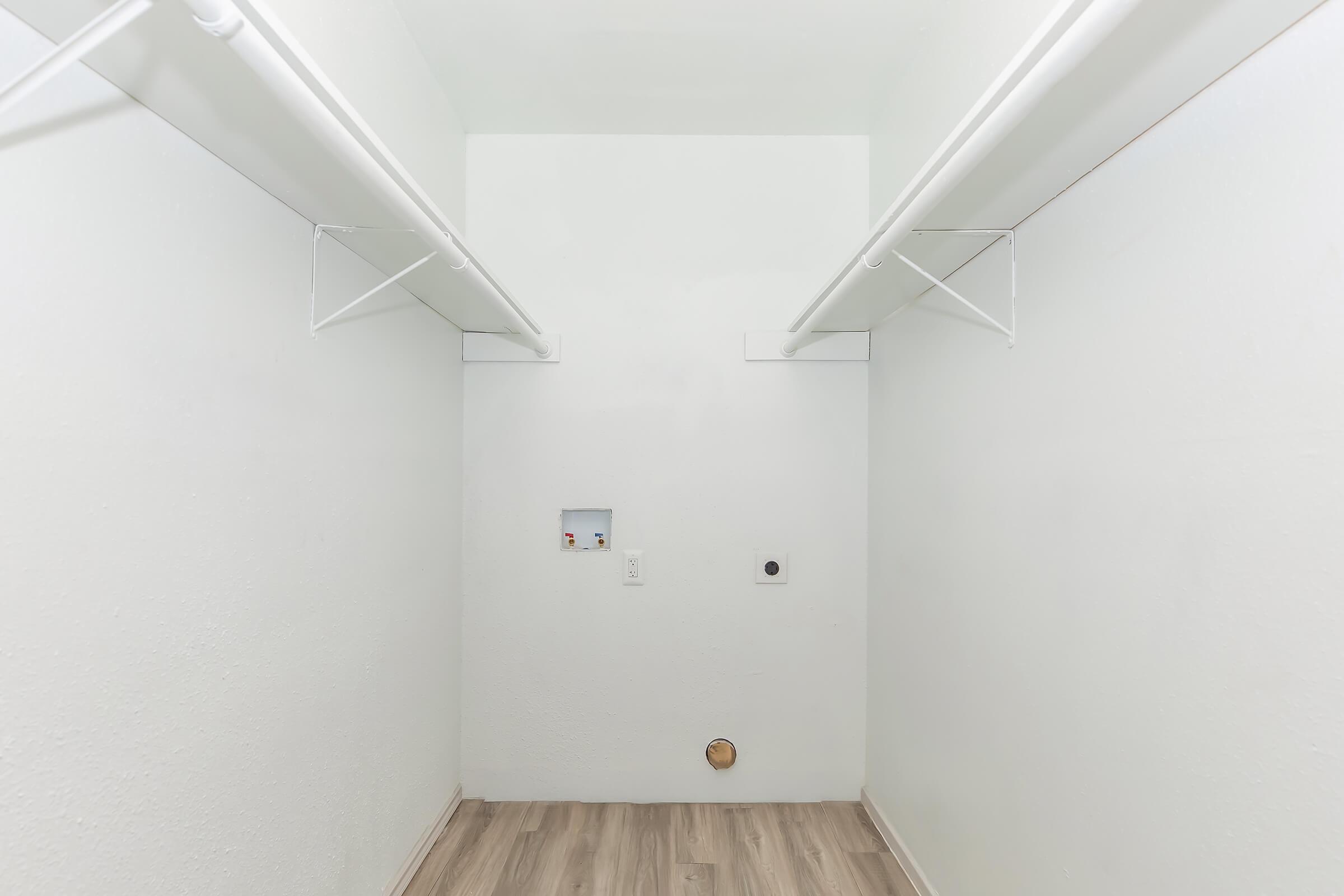 Empty room with white walls and two hanging clothes rods on opposite sides. The floor is light-colored wood. There is a small electrical panel and a circular wall opening on one side, indicating it may serve as a laundry or storage space. The room is well-lit and spacious with minimal decor.