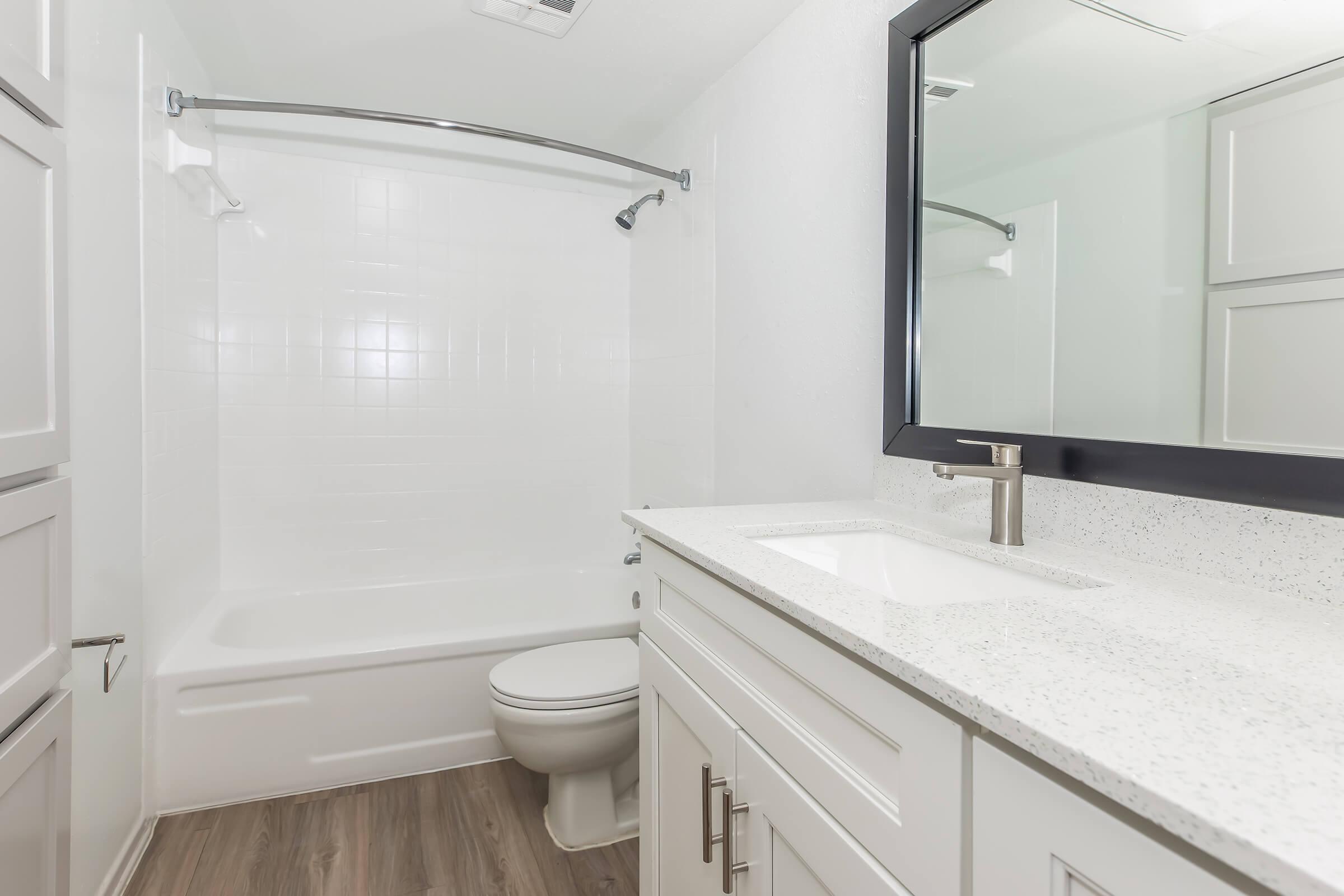A clean, modern bathroom featuring a white bathtub with a shower, a sleek white vanity with a sink, and a large mirror. The walls are painted light, and the floor has a wood-like appearance, creating a fresh and inviting space.