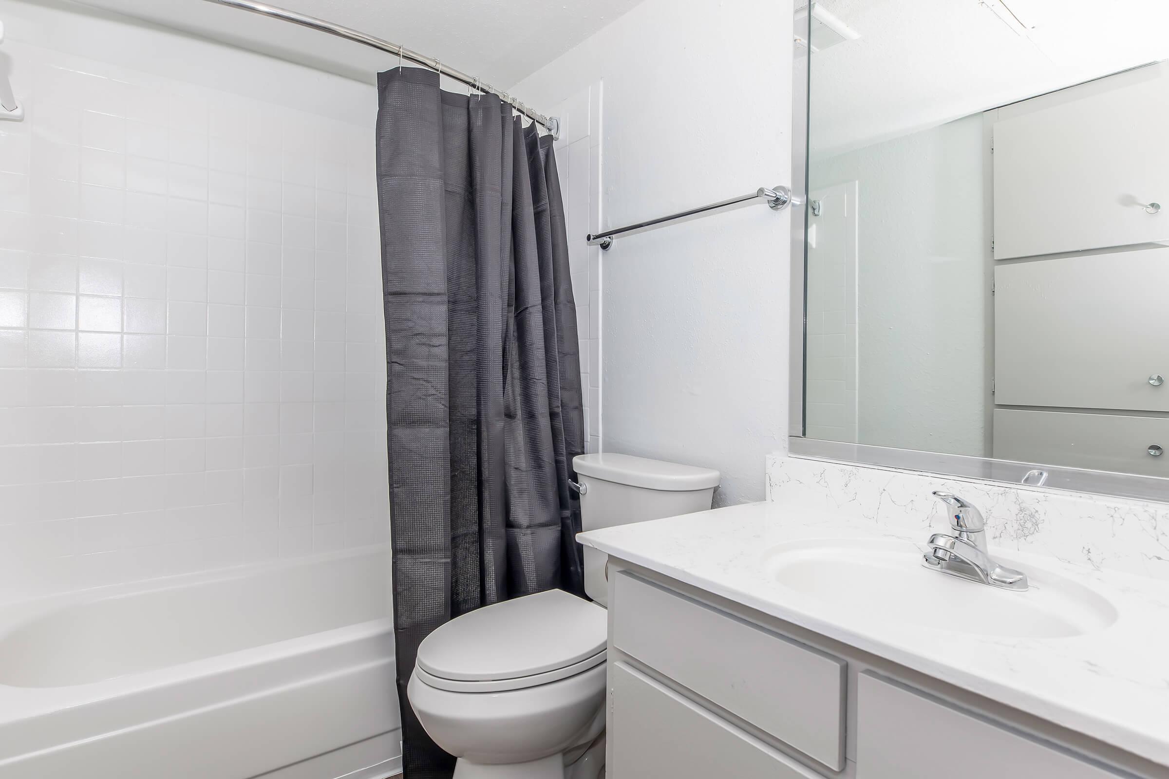 A clean bathroom featuring a bathtub with a shower curtain, a toilet, and a sink with a marble countertop. The walls are white, and there is a large mirror reflecting the room. Towel racks and storage cabinets are also visible.