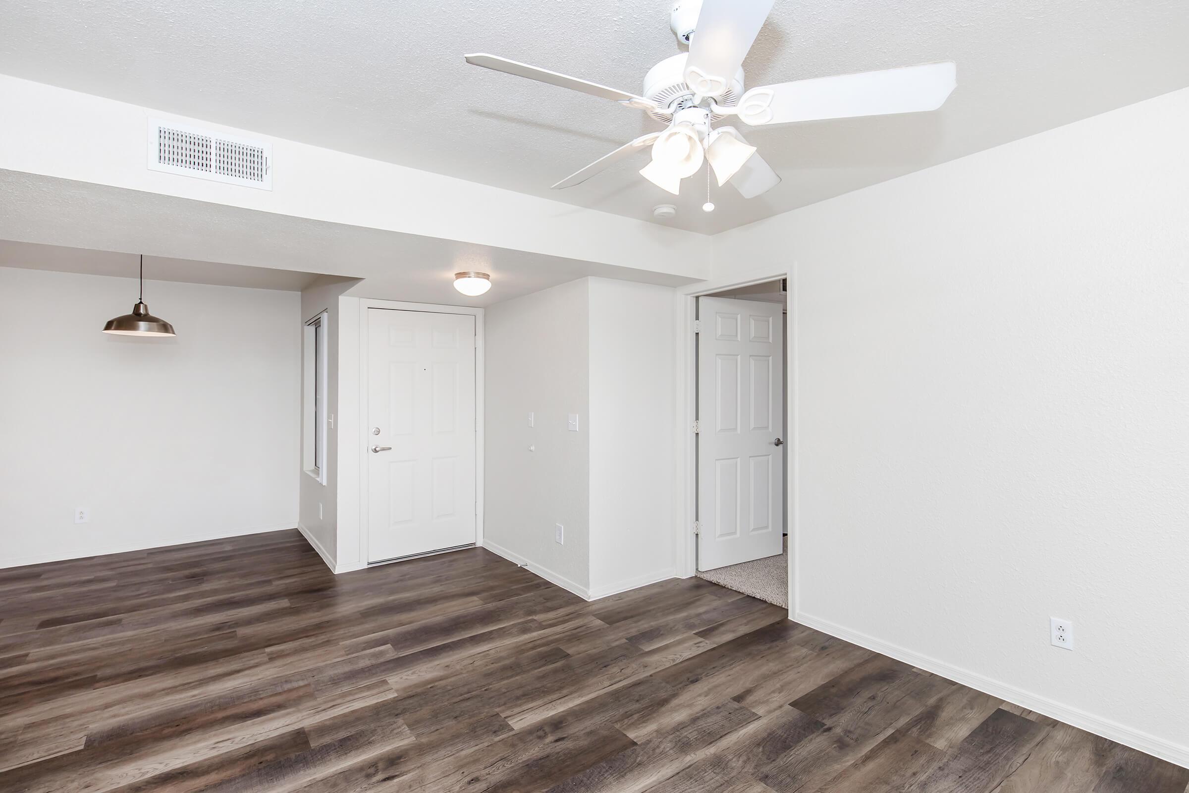 Interior of a modern apartment showing a spacious living area with wood-style flooring, white walls, and a ceiling fan. There is a door leading outside, a light fixture hanging above, and an adjacent doorway that opens into another room. The space is well-lit and designed for comfort.