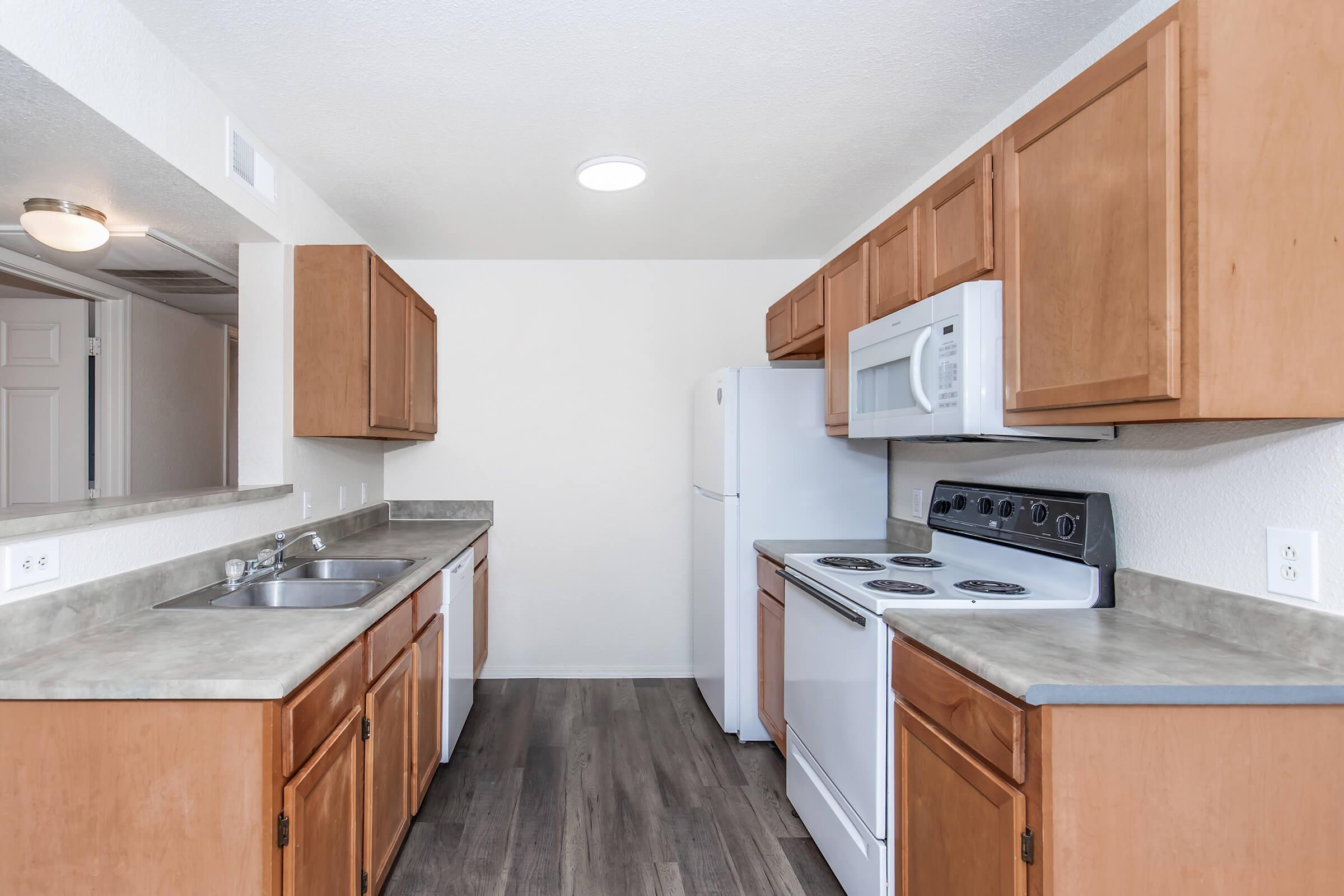 Modern kitchen featuring wooden cabinets, a white refrigerator, and an oven with a stovetop. The countertops are gray, and there is a double sink. The walls are painted white, and the space has a bright overhead light. The floor has a wood-like finish.
