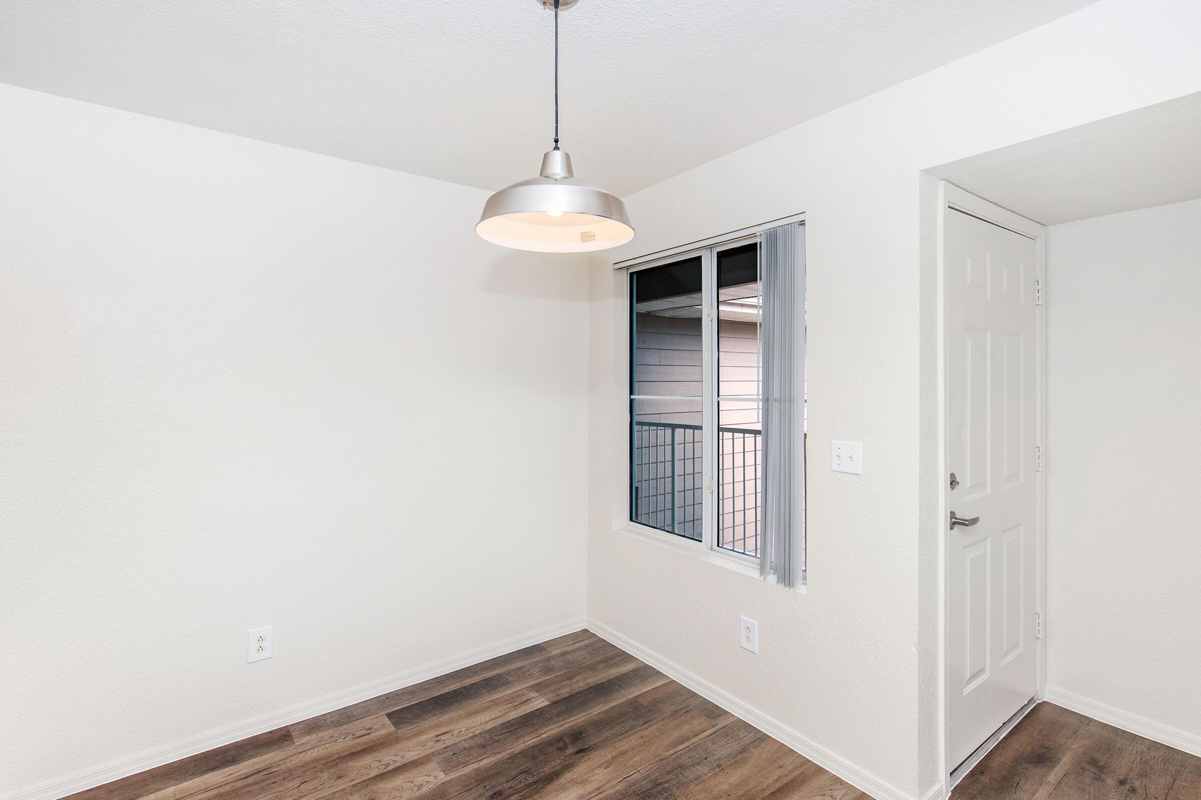 A well-lit, empty room featuring light-colored walls, a pendant light fixture, and a window with blinds. The flooring is dark wood, and there is a door on the right side leading to another space. The overall atmosphere is clean and minimalist, suitable for various purposes.