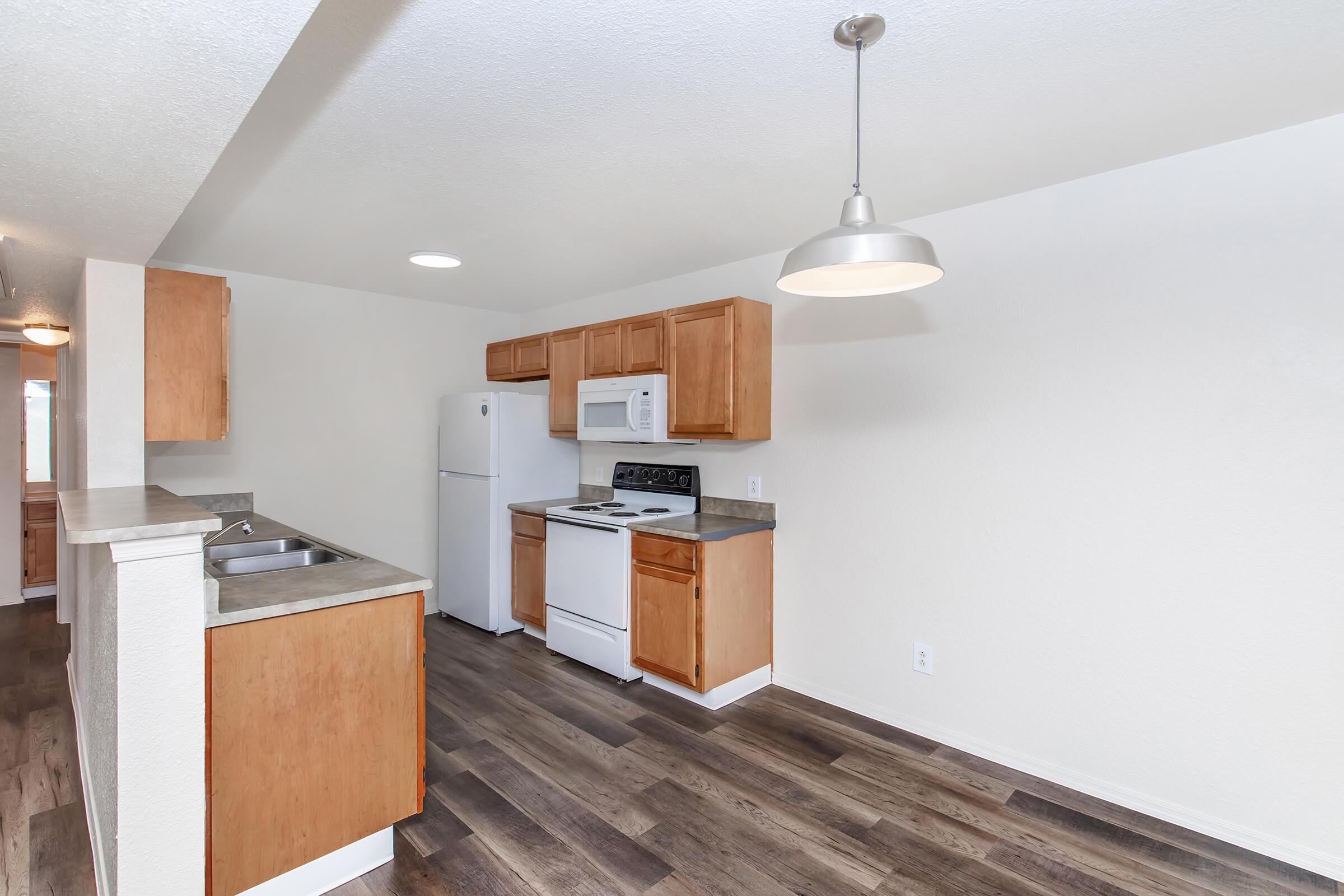 A spacious kitchen featuring wooden cabinets, a white refrigerator, a white microwave, and a gas stove. The room has a neutral-colored wall and ceiling lights, along with a pendant light above the island counter, and a view of the living area with wooden flooring.