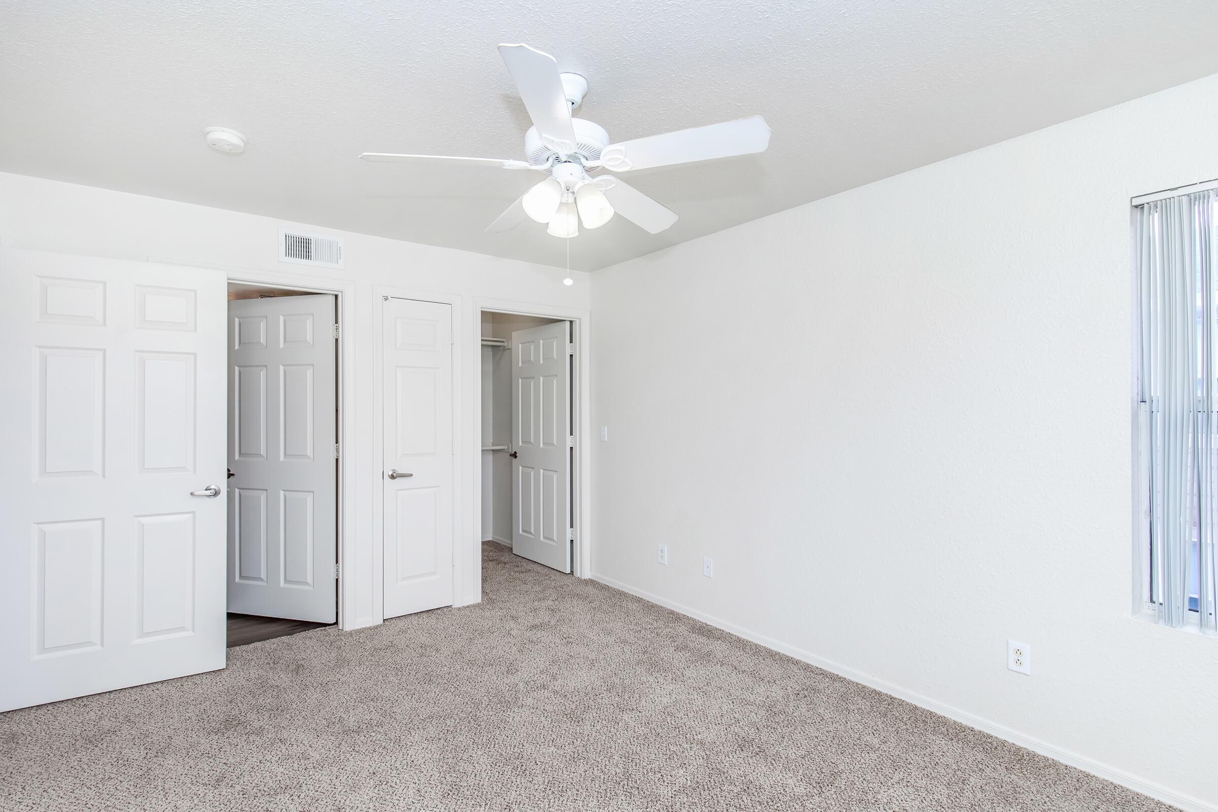 A well-lit room with light-colored walls and carpeted flooring. It features a ceiling fan with three lights, two closet doors on the left, and a window with vertical blinds on the right. The space is empty and unfurnished, providing a clean and open atmosphere.