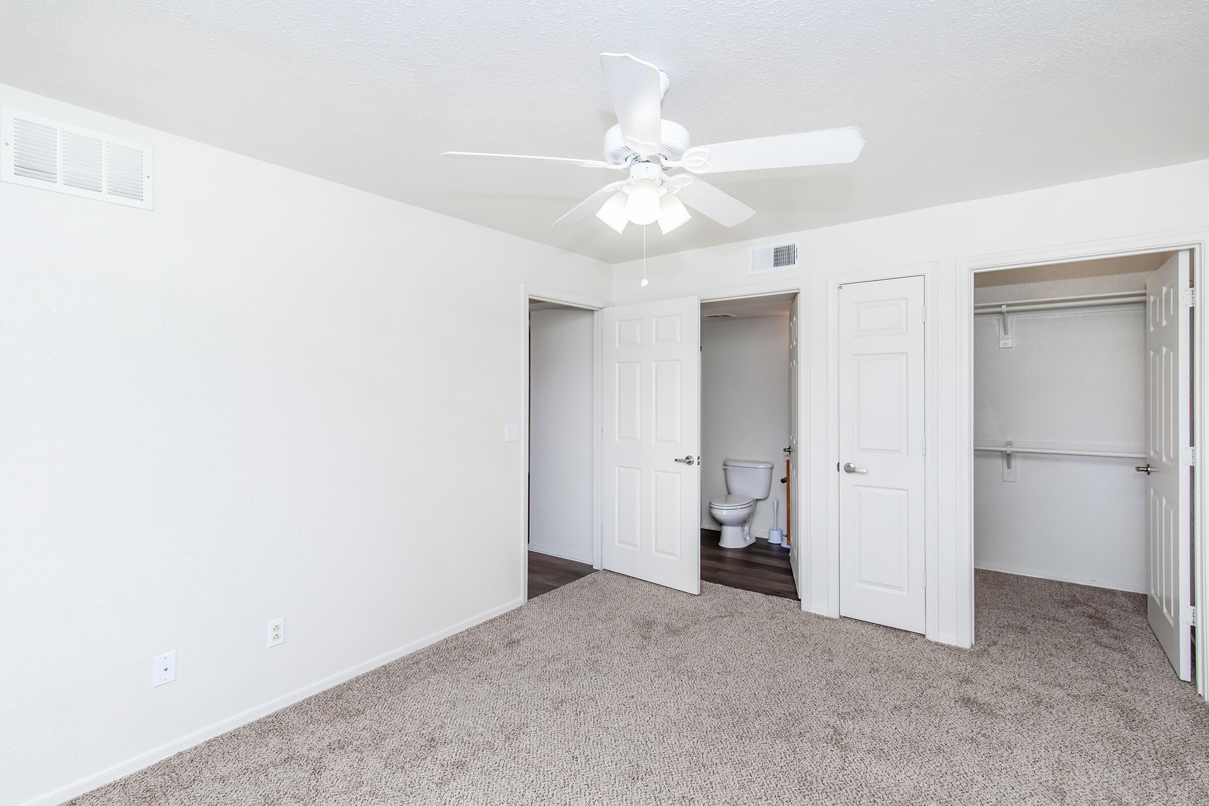 A bright, minimalistic bedroom featuring light-colored walls and carpet. There is a ceiling fan and two closed doors, one leading to a bathroom with a toilet and the other to a closet. The room is well-lit and clean, creating a spacious feel.