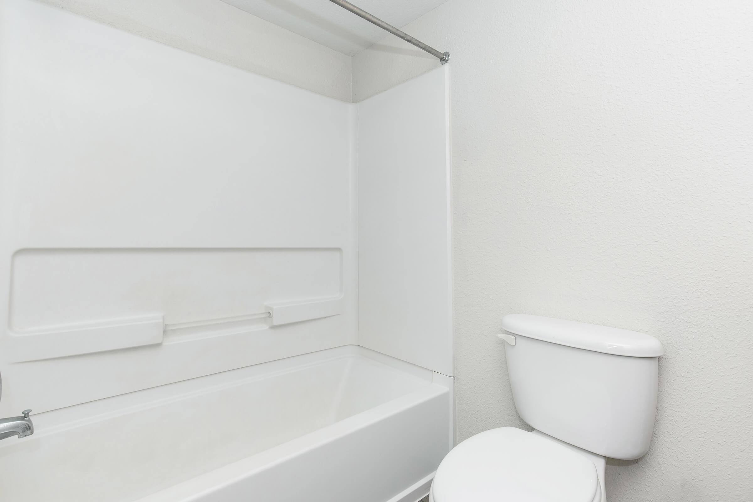 A clean and simple bathroom featuring a white bathtub, a shower rod, and a toilet. The walls are painted light colors, creating a bright and spacious appearance. The bathroom fixtures are basic and modern, with a minimalistic design.