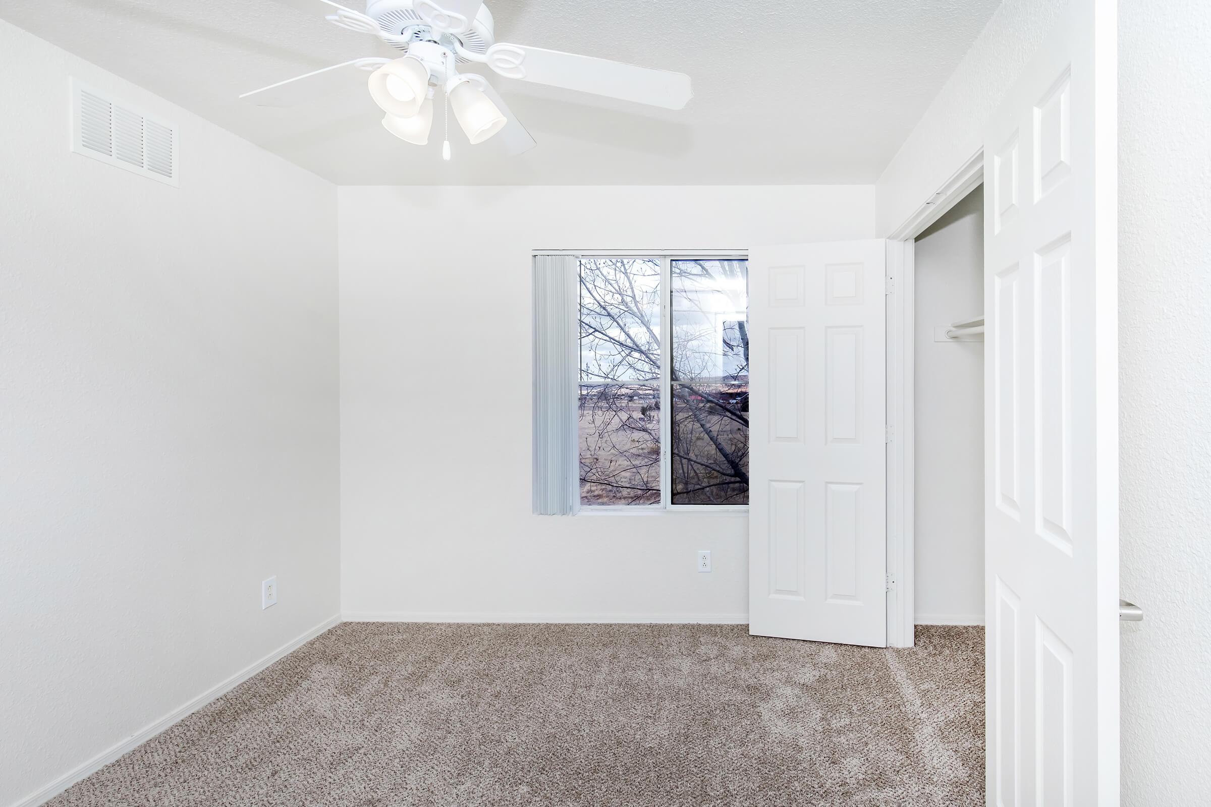 A well-lit, empty bedroom featuring light-colored walls, a ceiling fan with five blades, and plush carpet. A large window with sheer blinds allows natural light in, and the closet door is open, revealing a simple closet space. The room has a clean, minimalist appearance with no furniture.