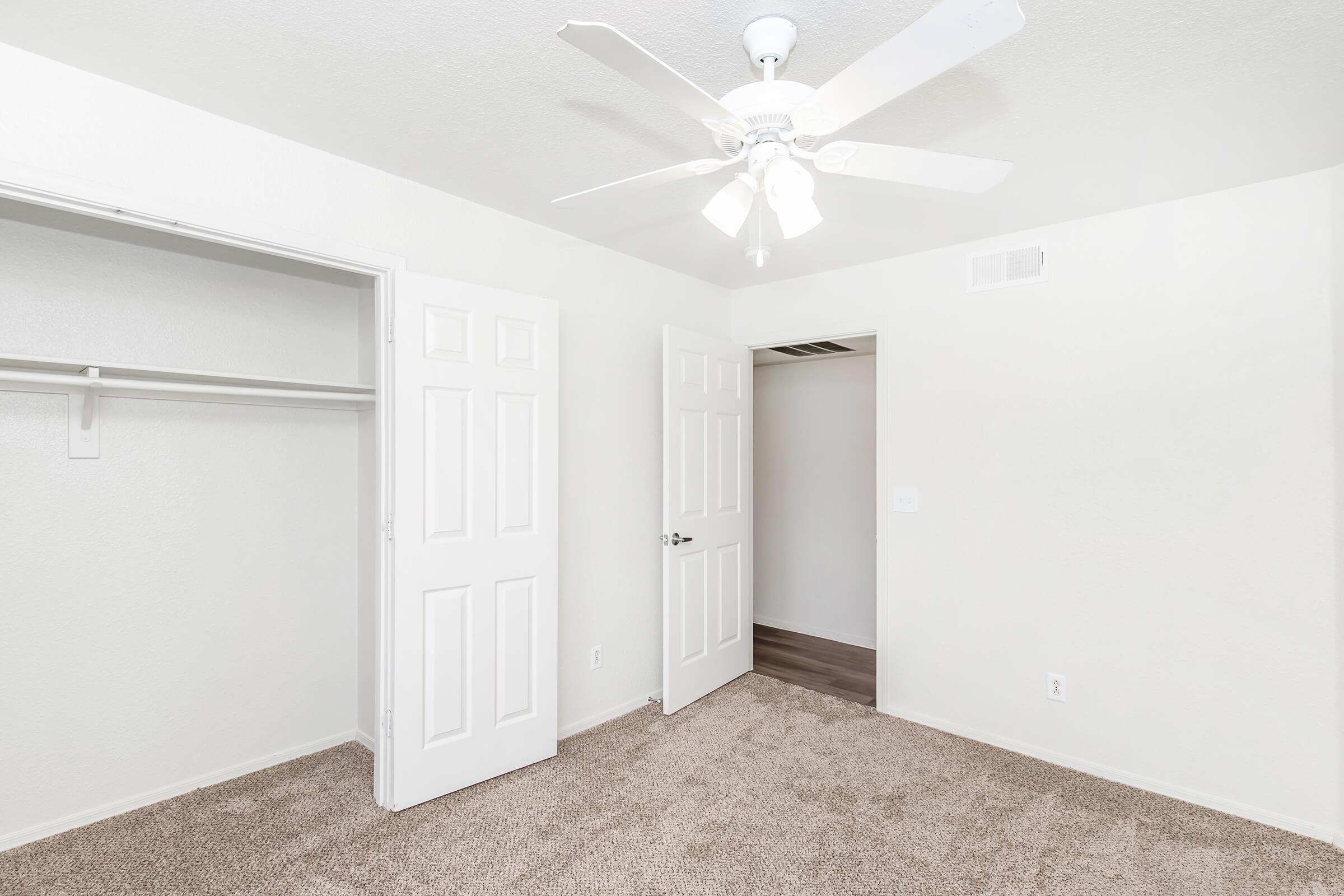 A well-lit, empty bedroom featuring light beige carpet, white walls, and a ceiling fan. There are two doors: one leading to a closet with a shelf and the other to an adjacent room. The space is clean and minimalistic, suitable for various décor styles.