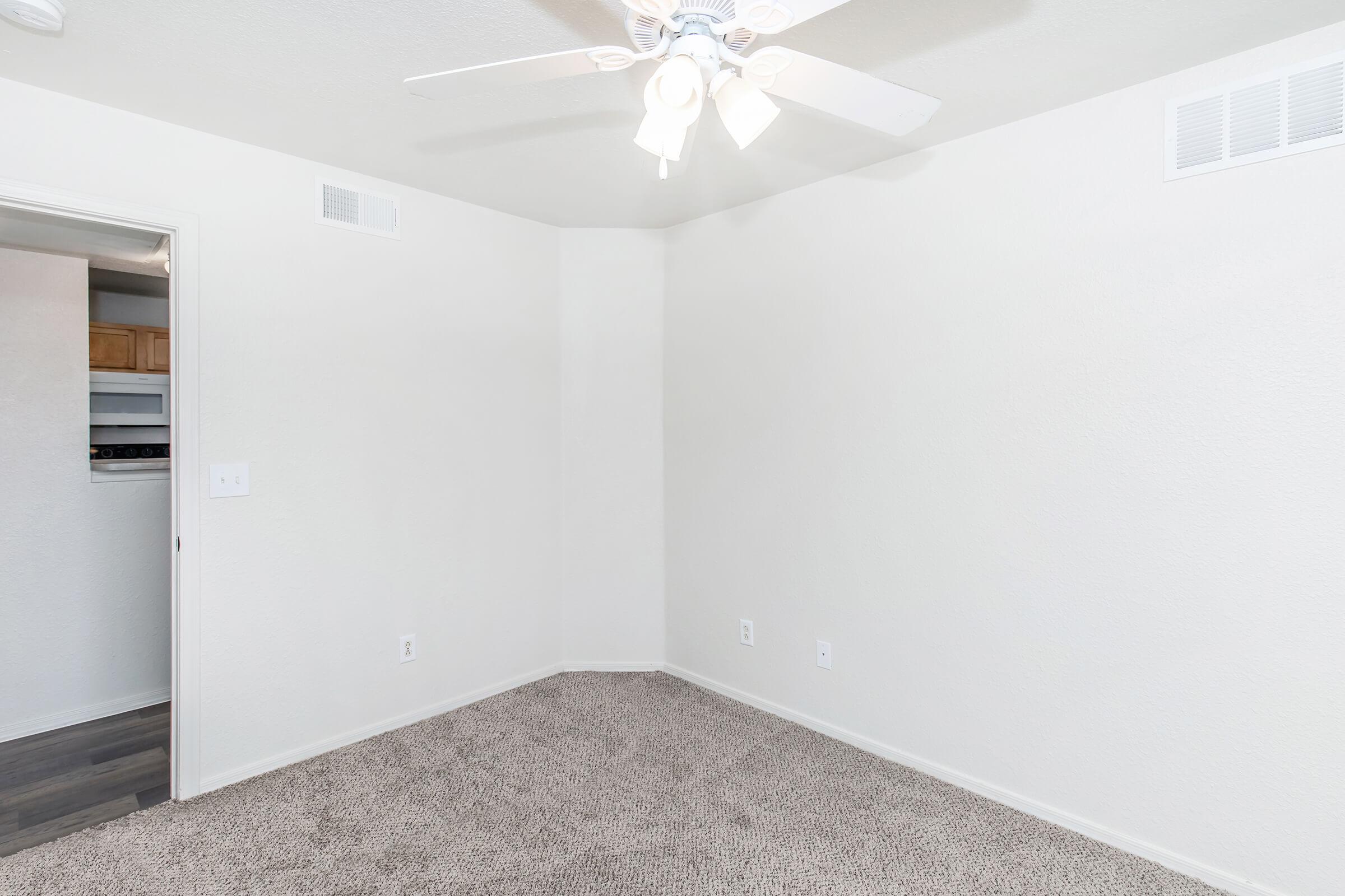 An empty room with light-colored walls and a ceiling fan. The floor is covered in beige carpet, and there is a doorway leading to another room with a laundry area visible. The room has no furniture, creating a spacious and minimalist feel.