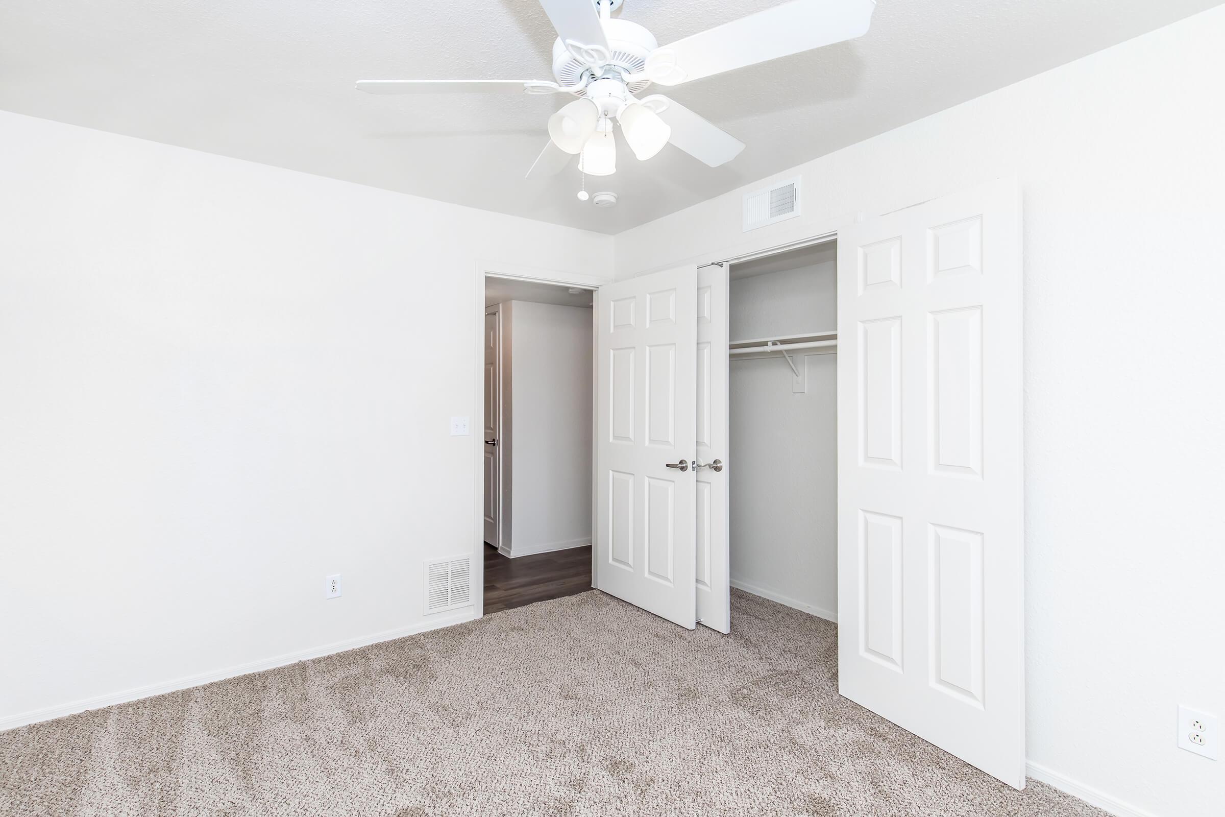 A clean, well-lit bedroom featuring beige carpet, white walls, and a ceiling fan. Two doors lead to a closet space and an adjacent room. The room has a spacious, uncluttered feel, emphasizing a minimalistic design.