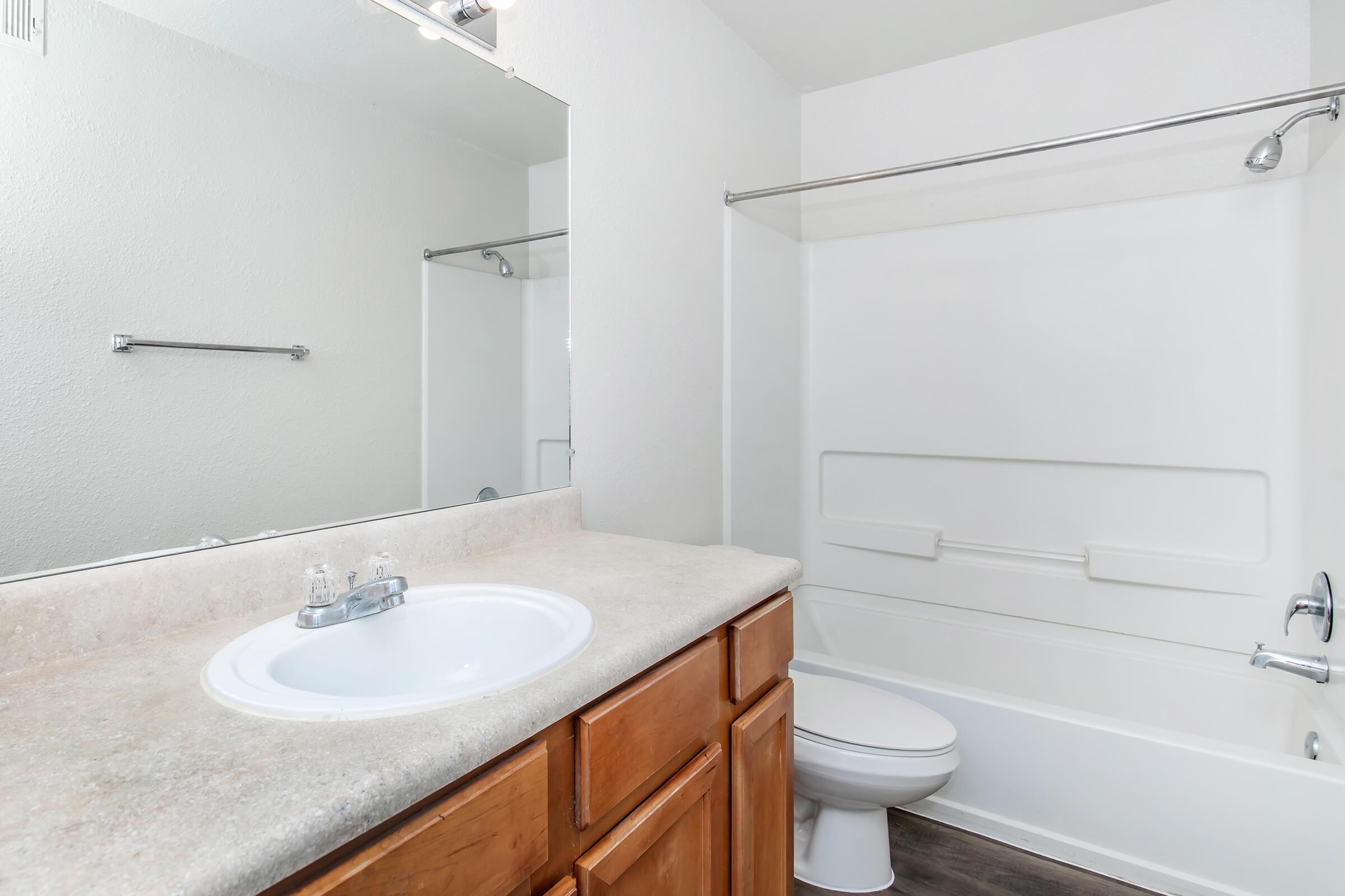 A clean, modern bathroom featuring a white bathtub with a shower curtain, a single sink with a light wood vanity, and a large mirror above the sink. The walls are painted in a light color, and the floor is dark, providing a contrast to the overall bright ambiance.