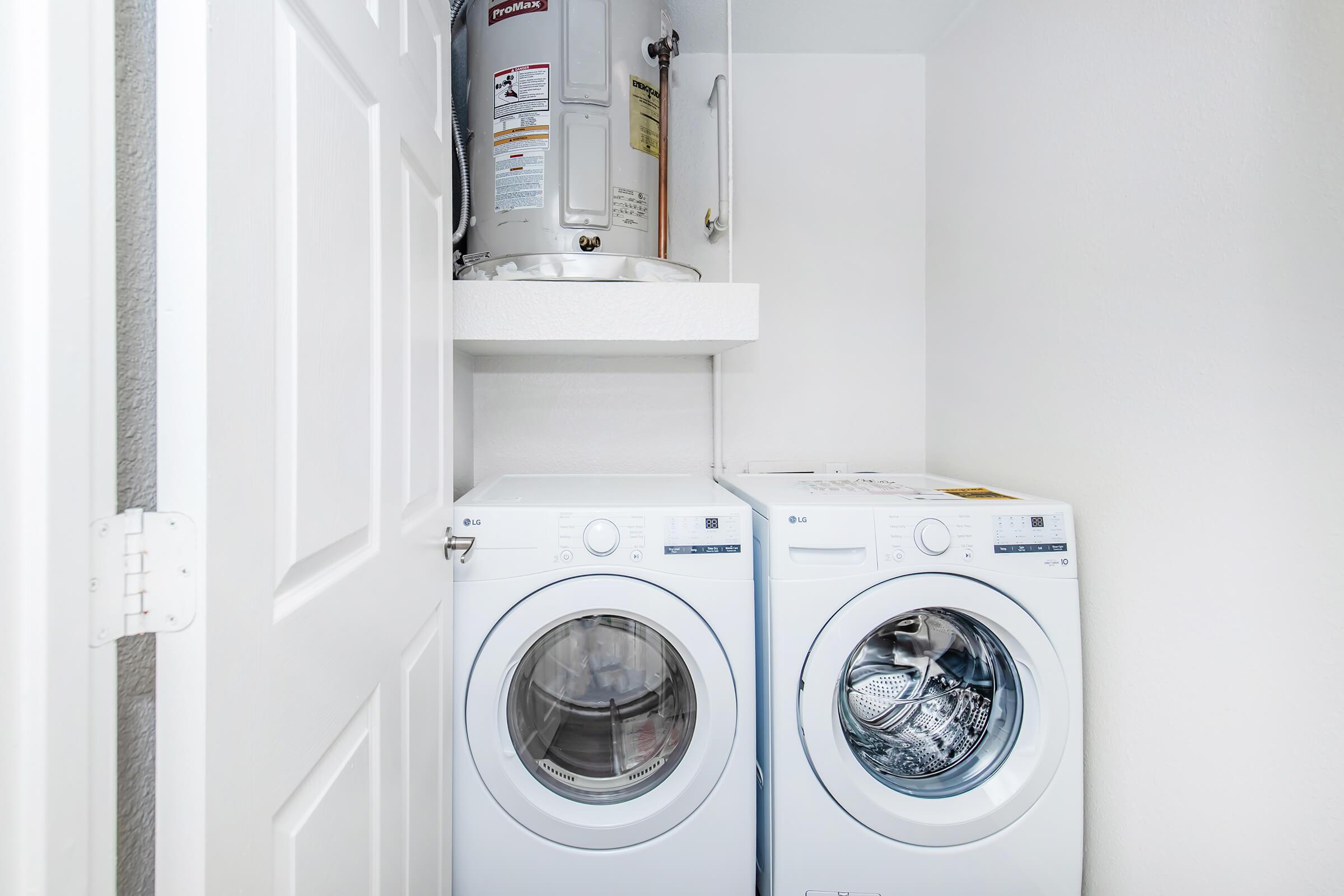A clean and minimalist laundry area featuring a stacked washer and dryer in white. Above them is a shelf with a water heater. The walls are painted light, contributing to a bright and organized space. The door is partially open, revealing the compact laundry setup.