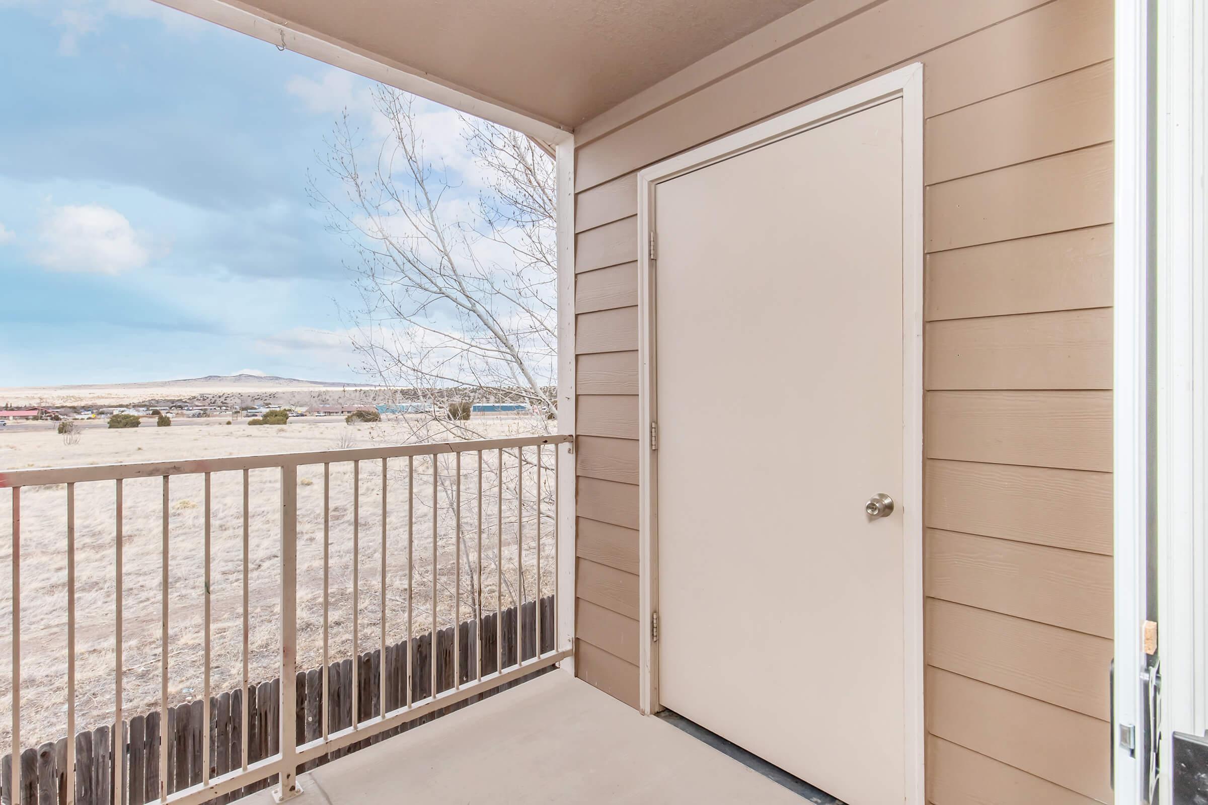 View of a balcony with a white door and beige siding. The area outside has a grassy field and a distant mountain range under a partly cloudy sky. The railings are simple and the setting appears peaceful and quiet.