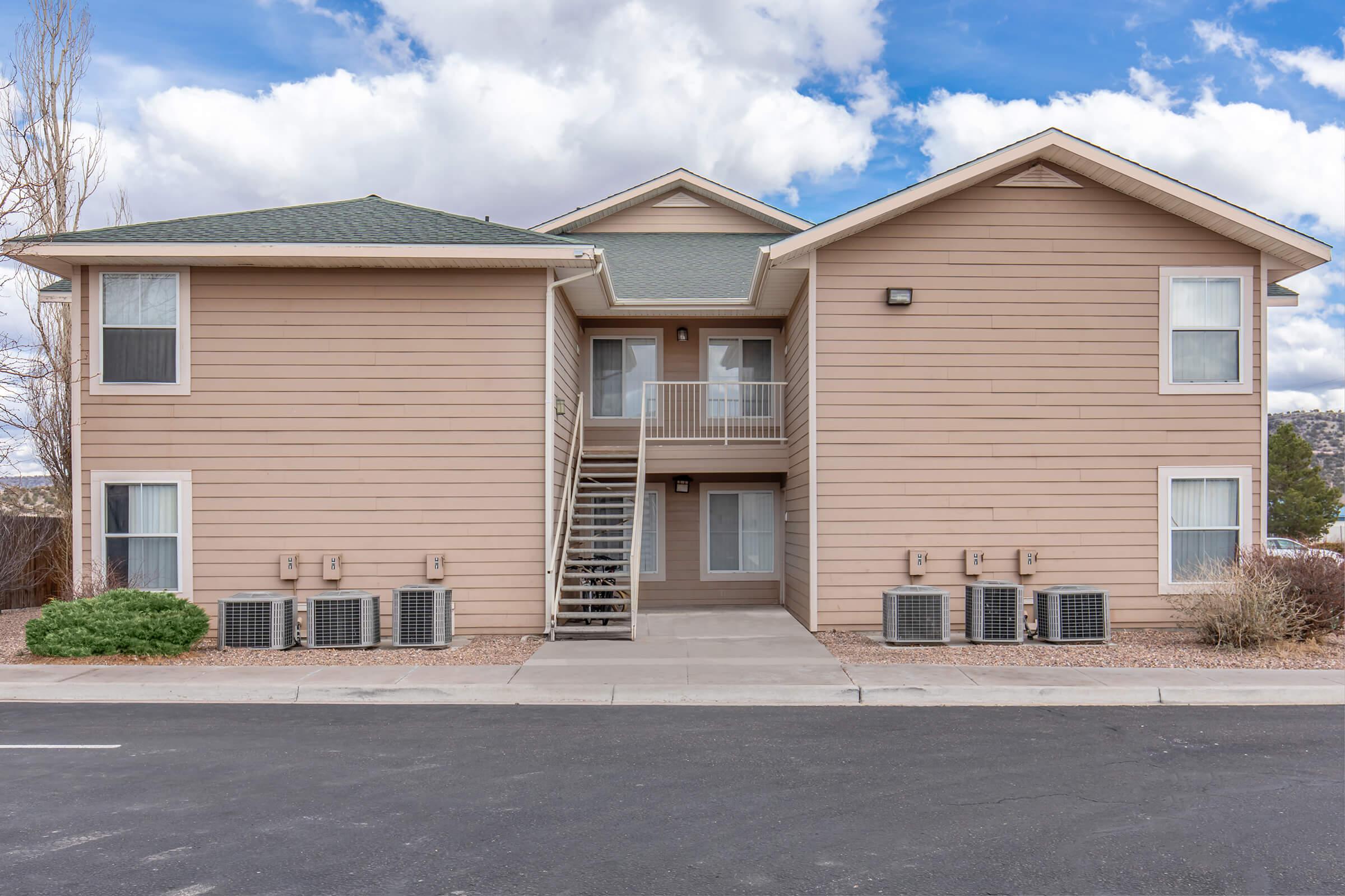 A two-story beige apartment building with a green roof. It features a staircase leading to the second floor, multiple air conditioning units on the ground level, and landscaped shrubs. The sky above is partly cloudy with blue spots peeking through.