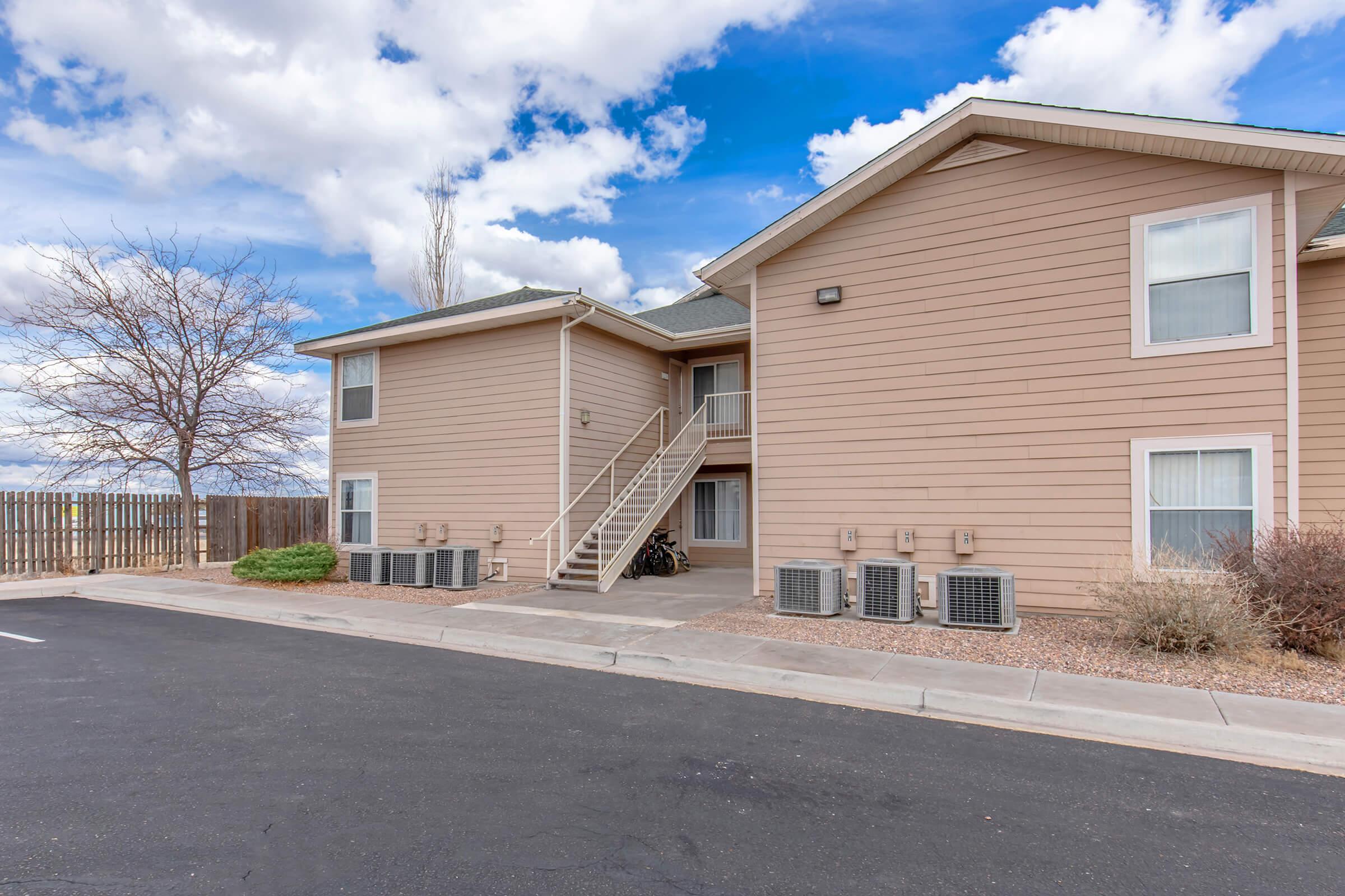 Exterior view of a two-story residential building with a light brown facade. Stairs lead up to the second floor. There are several air conditioning units along the ground level, and a bike is parked near the entrance. The sky is partly cloudy, and there is a barren tree nearby.