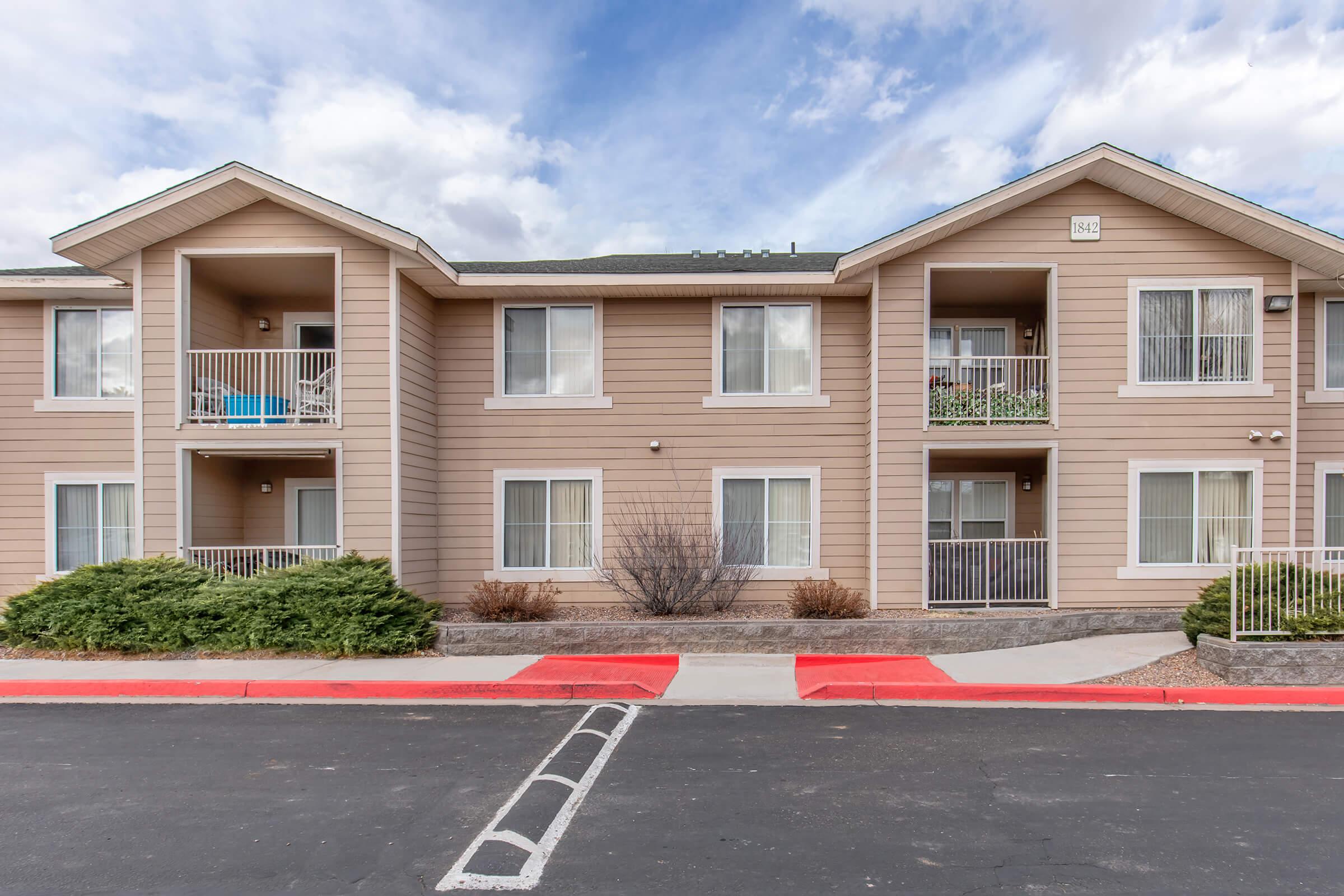 Exterior view of a multi-story apartment building with a light brown facade. The building features balconies with potted plants, windows with white frames, and landscaped shrubs in front. The pavement is marked with a red pedestrian pathway leading to the entrance. The sky is partly cloudy.