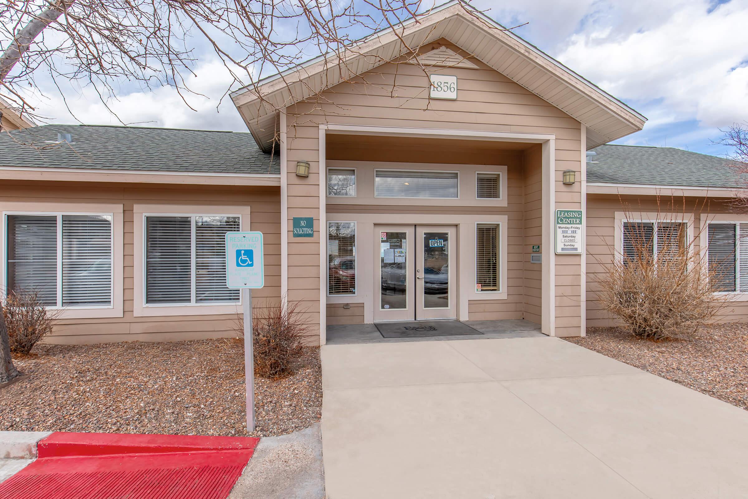Single-story building with a wide entrance featuring large windows. The facade is light brown with white trim. There is a sign indicating "Leasing Office" next to the entrance. A blue parking sign for accessibility is visible in front of the building. The surrounding area includes gravel landscaping and a cloudy sky.