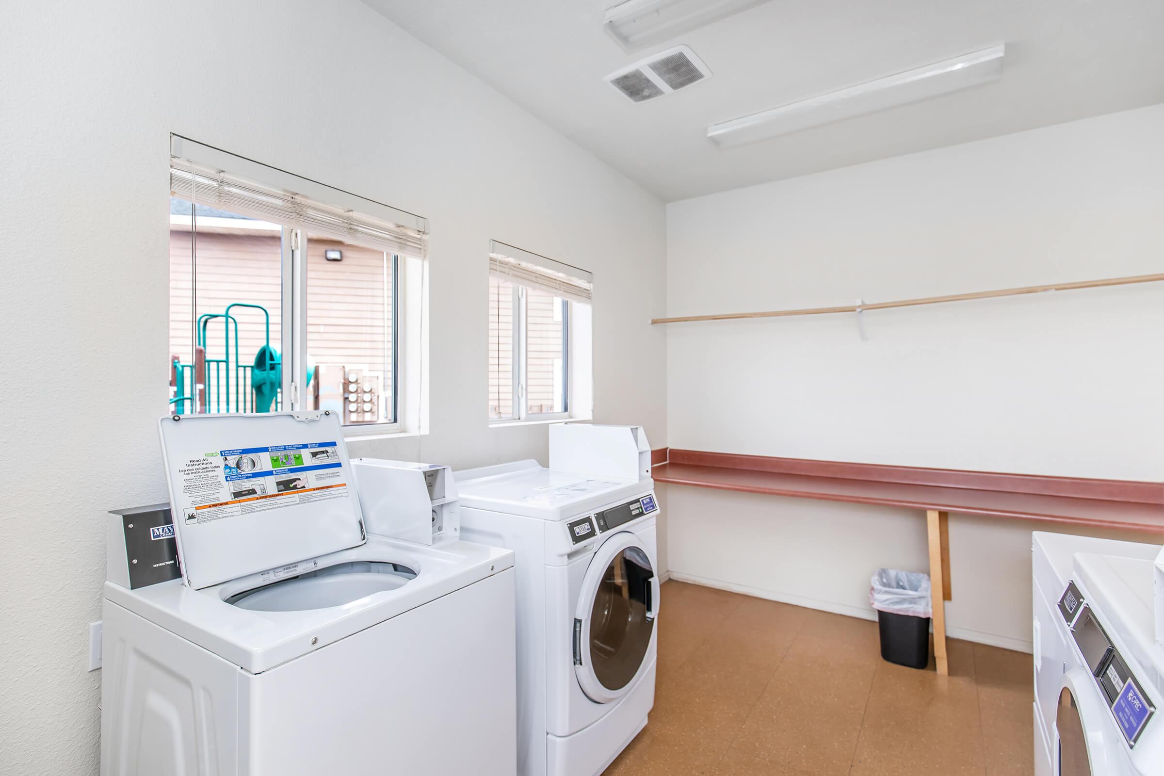 A clean and bright laundry room featuring two white washing machines. There are large windows allowing natural light, a red countertop along one wall, a trash can, and a green playground visible outside through the windows. The walls are painted white, enhancing the room's spacious feel.