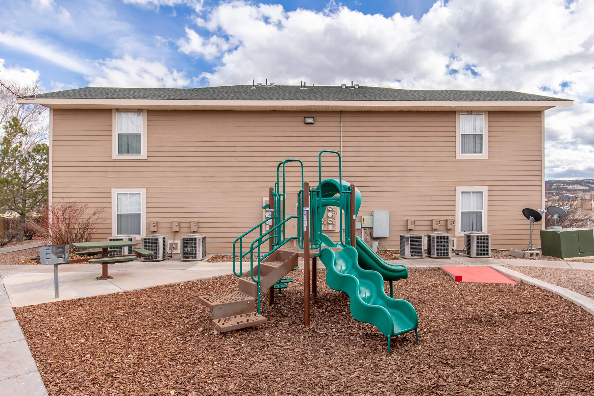 A playground area featuring a green slide and climbing structure, surrounded by mulch. In the background, a beige building with multiple windows and air conditioning units. The sky is partly cloudy, contributing to a bright outdoor atmosphere.