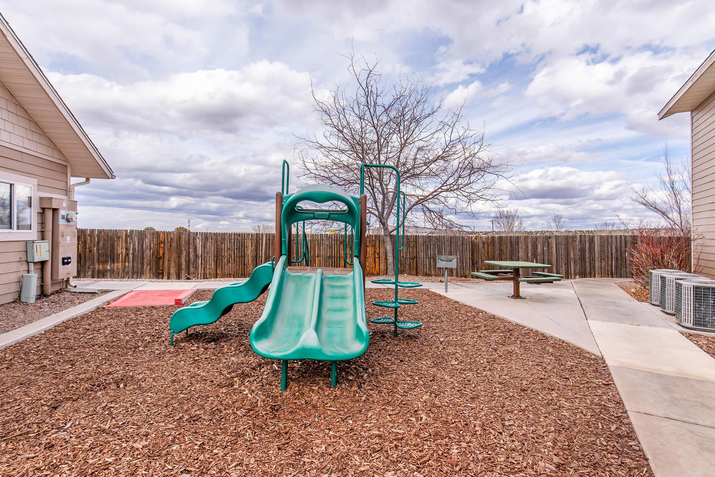 A playground featuring a green slide and climbing structure situated on a mulch-based surface. In the background, there is a wooden fence and cloudy skies, with a bare tree nearby. A picnic table is visible at the right, along with air conditioning units near the buildings.