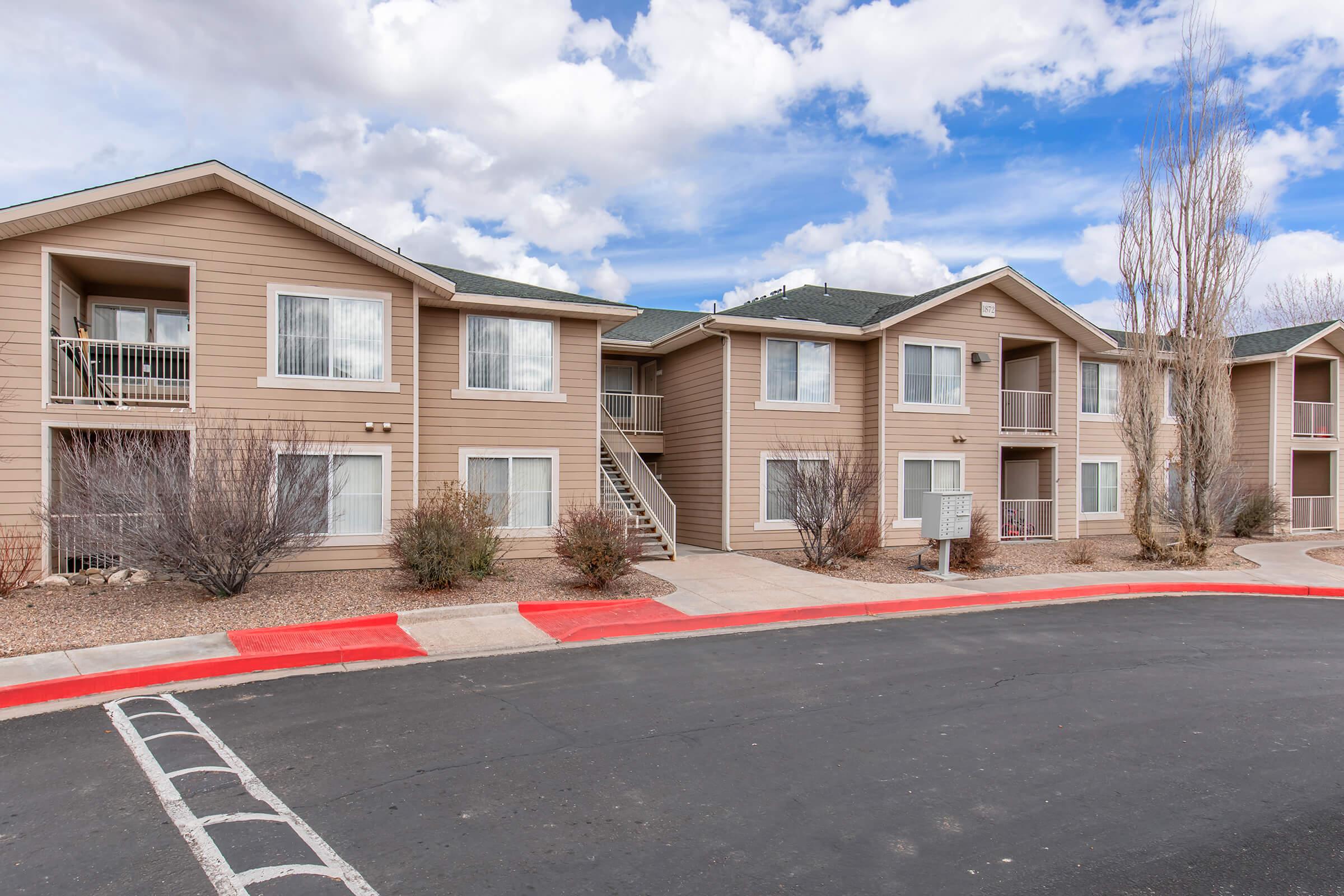 Two-story apartment building with beige siding and multiple balconies, surrounded by low shrubs and gravel landscaping. The sky is partly cloudy, and a red-painted curb lines the parking area in front of the apartments.