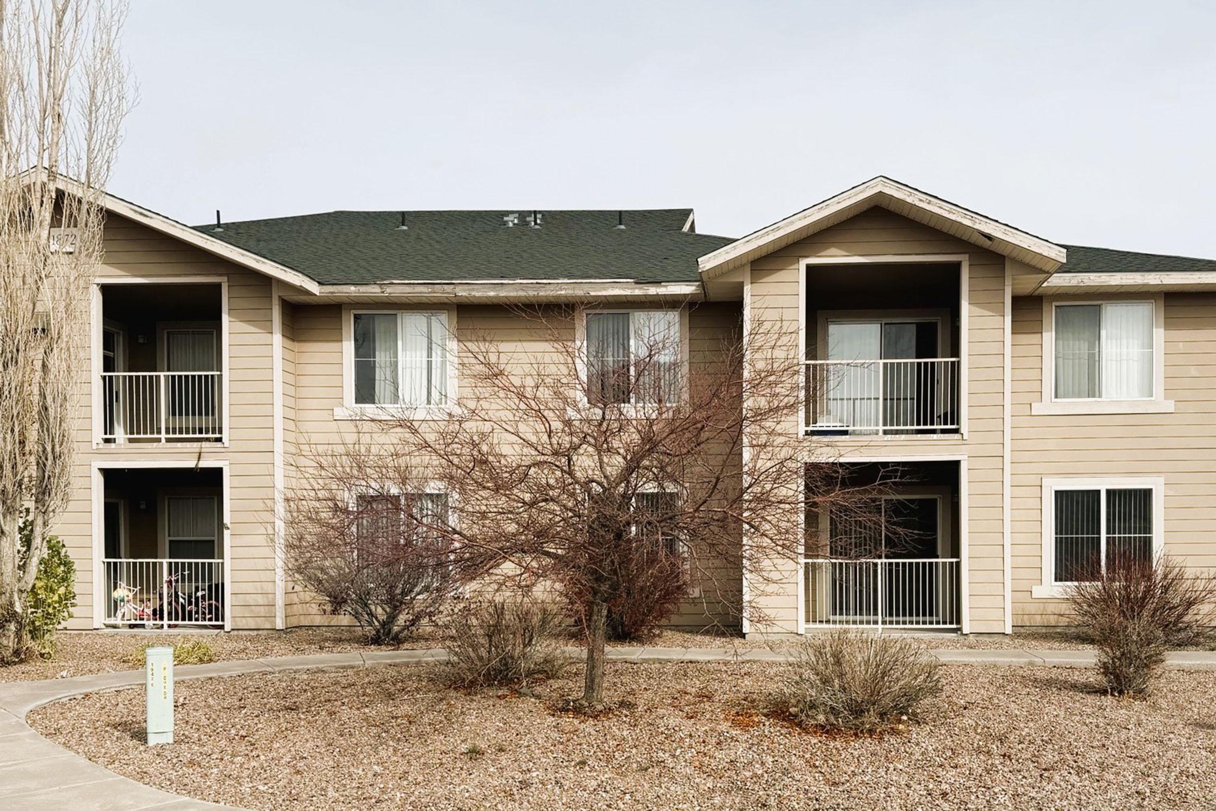 A two-story apartment building with multiple balconies, surrounded by a landscaped area featuring shrubs and small trees. The building has a light tan exterior with green accents. The sky is cloudy, giving a muted ambiance to the scene.