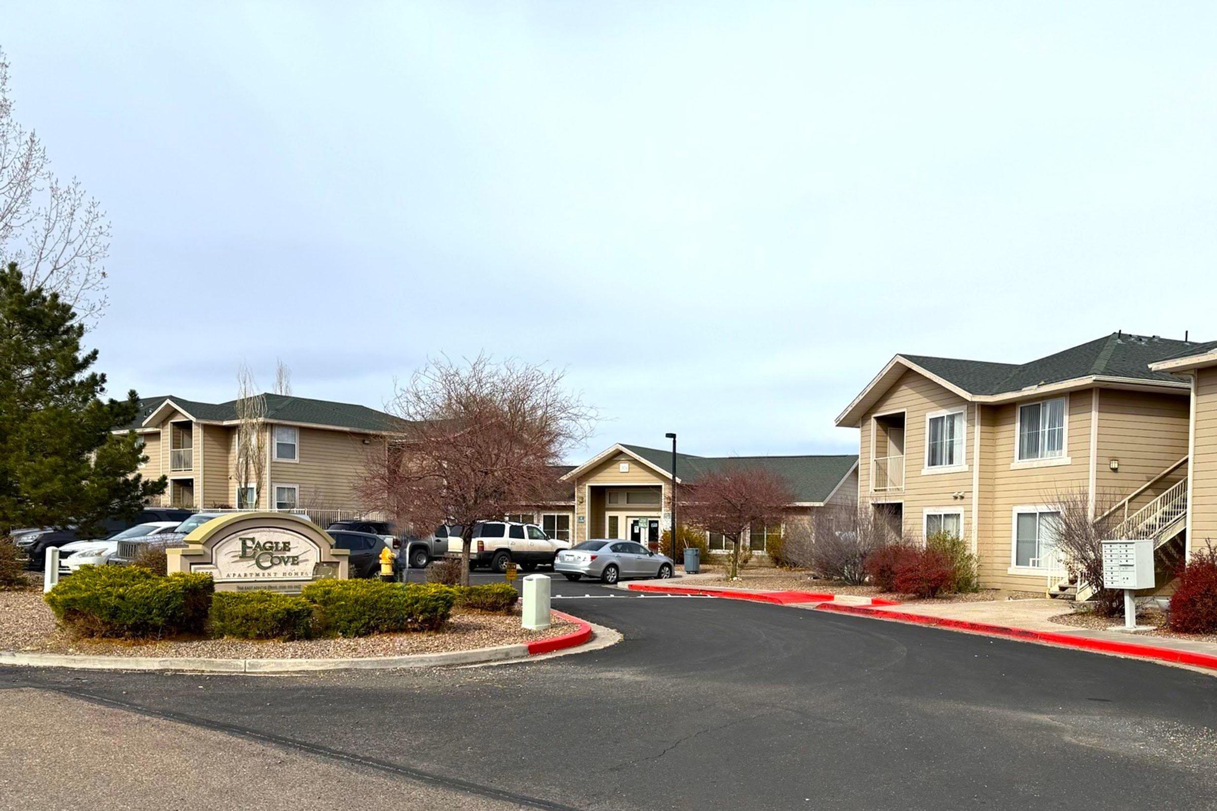 A view of a residential complex with beige multi-unit buildings, landscaped shrubs, and a red circular driveway. In the foreground, there is a sign for "Eagle Cove" and visible parking spaces. The sky is partly cloudy, creating a serene atmosphere.