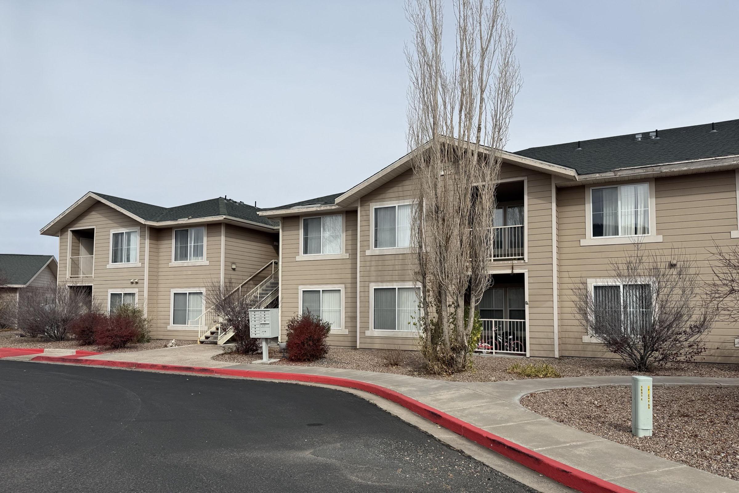 A tan apartment complex featuring multiple units with balconies, surrounded by small shrubs and trees. A curved driveway with a red curb leads to the building, and a sign is visible near the entrance. The sky is cloudy, creating a muted atmosphere.
