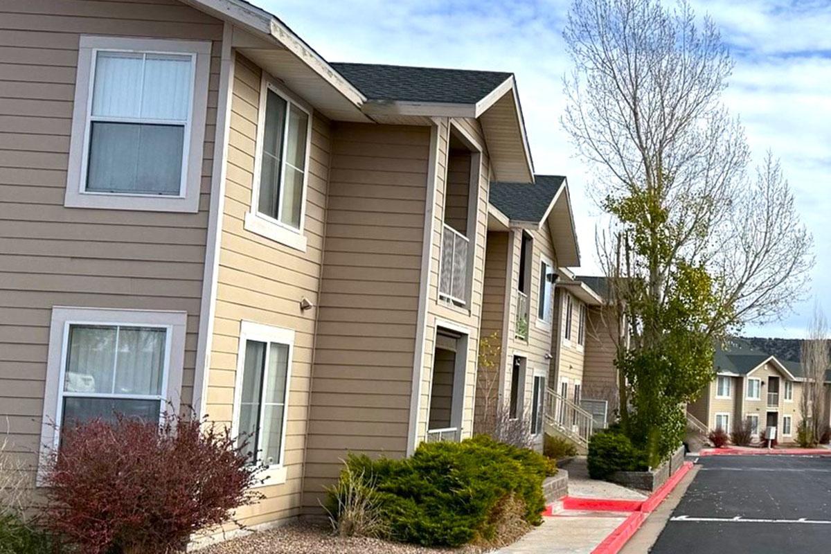 A row of multi-story apartment buildings with beige siding, a clear blue sky above, and well-maintained landscaping along the walkway. The buildings feature balconies and a mix of shrubs and trees in front, with a red-paved pathway visible on the side.