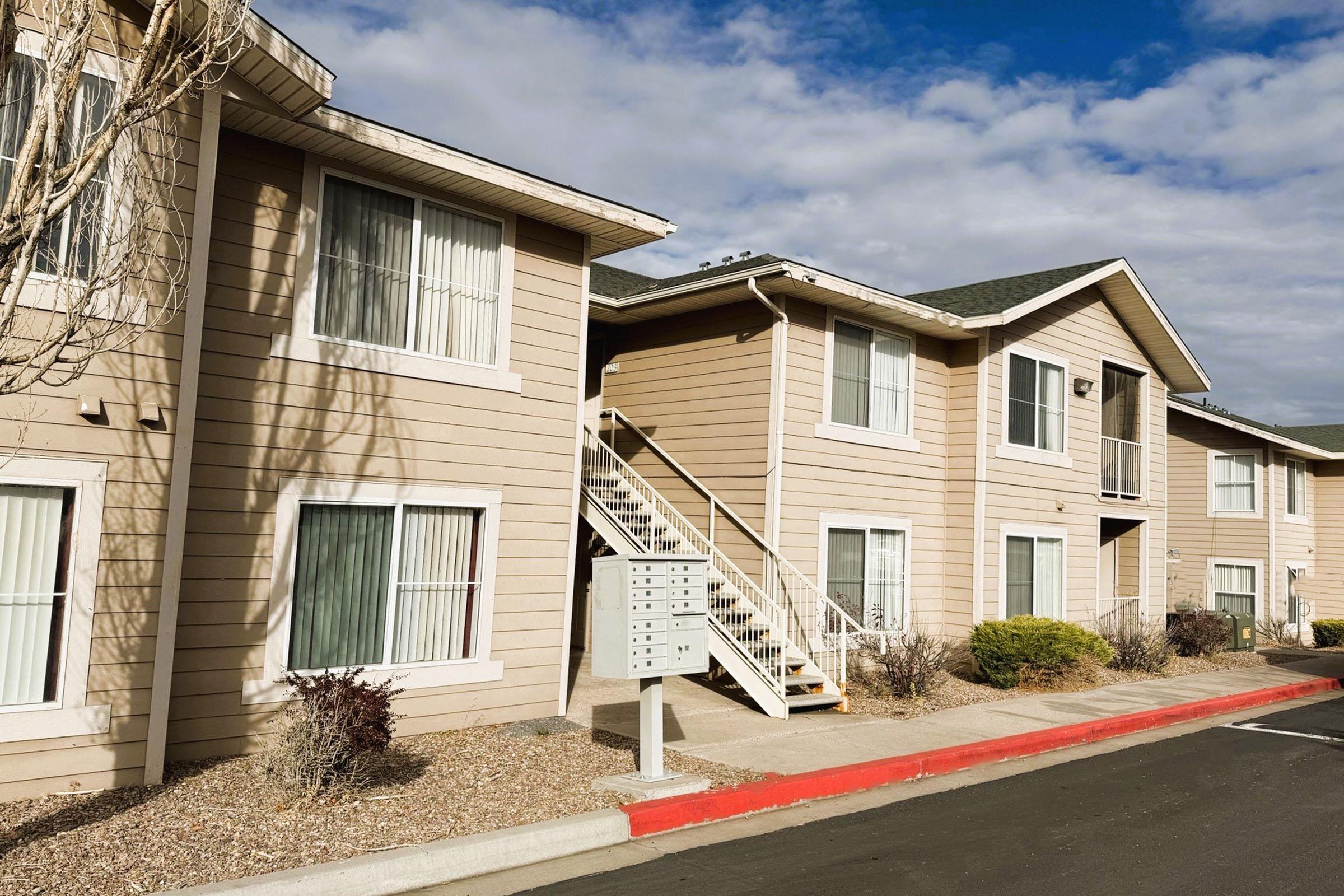 A residential apartment building with beige siding, featuring multiple windows and a staircase leading to the upper level. The area has light landscaping and a mailbox structure nearby. The sky is partly cloudy, creating a pleasant atmosphere.